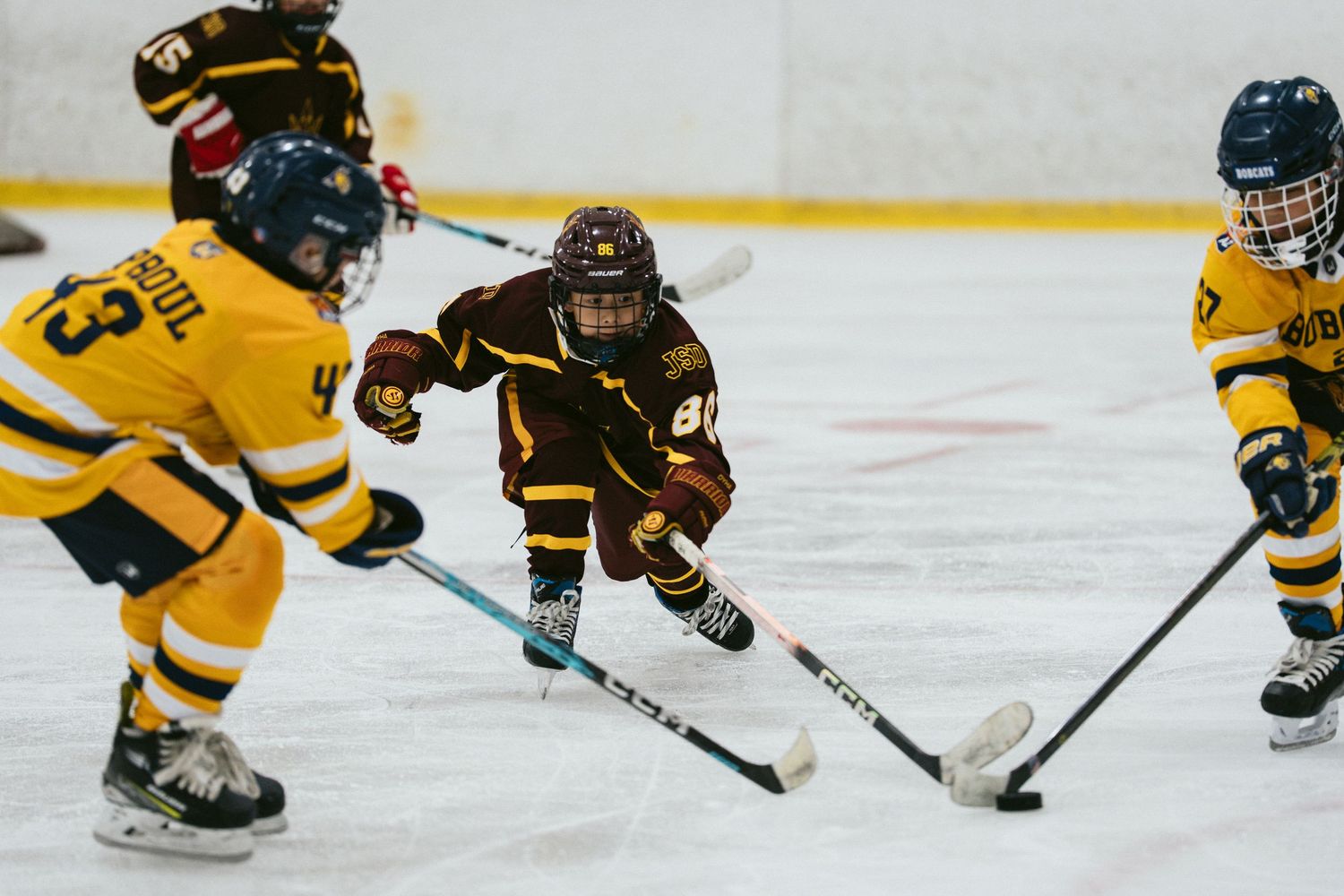Youth hockey players compete on ice, with a player in maroon jersey skating between two opponents in yellow uniforms.