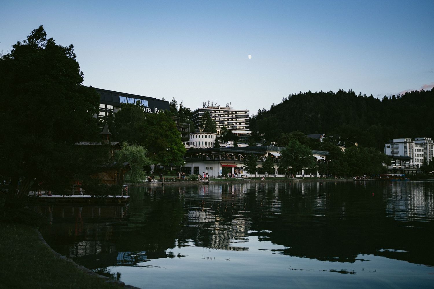 Tranquil lakeside town at dusk with reflections of buildings and trees on calm water under a crescent moon.