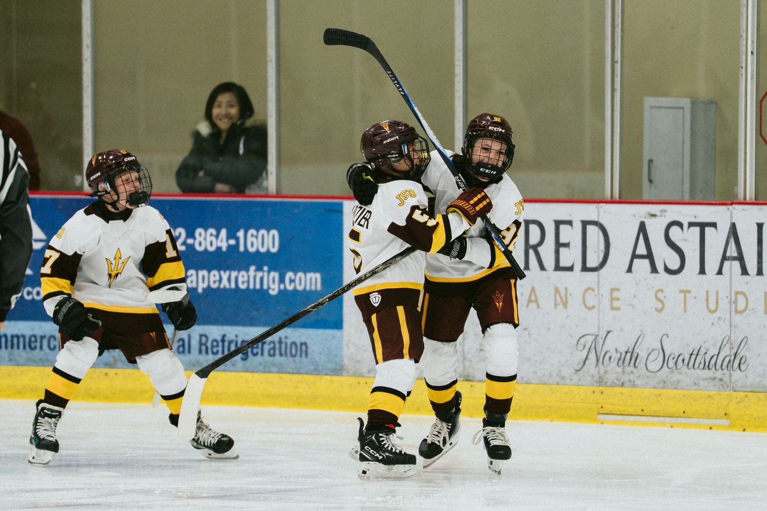 Youth hockey players in gold and white uniforms celebrate a goal on the ice rink during a game.