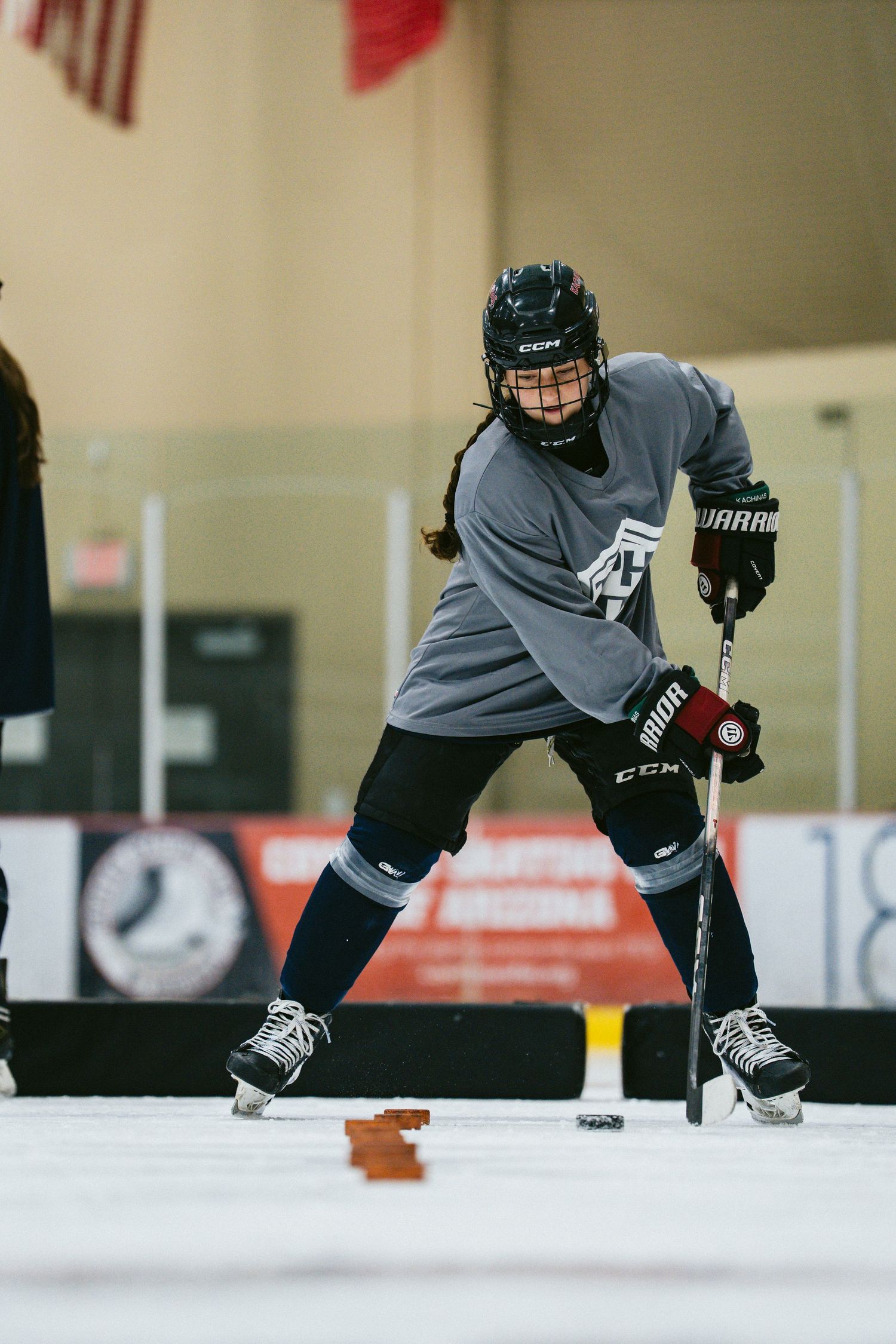 A young female ice hockey player in a gray CCM jersey and helmet handles a puck on an indoor rink.