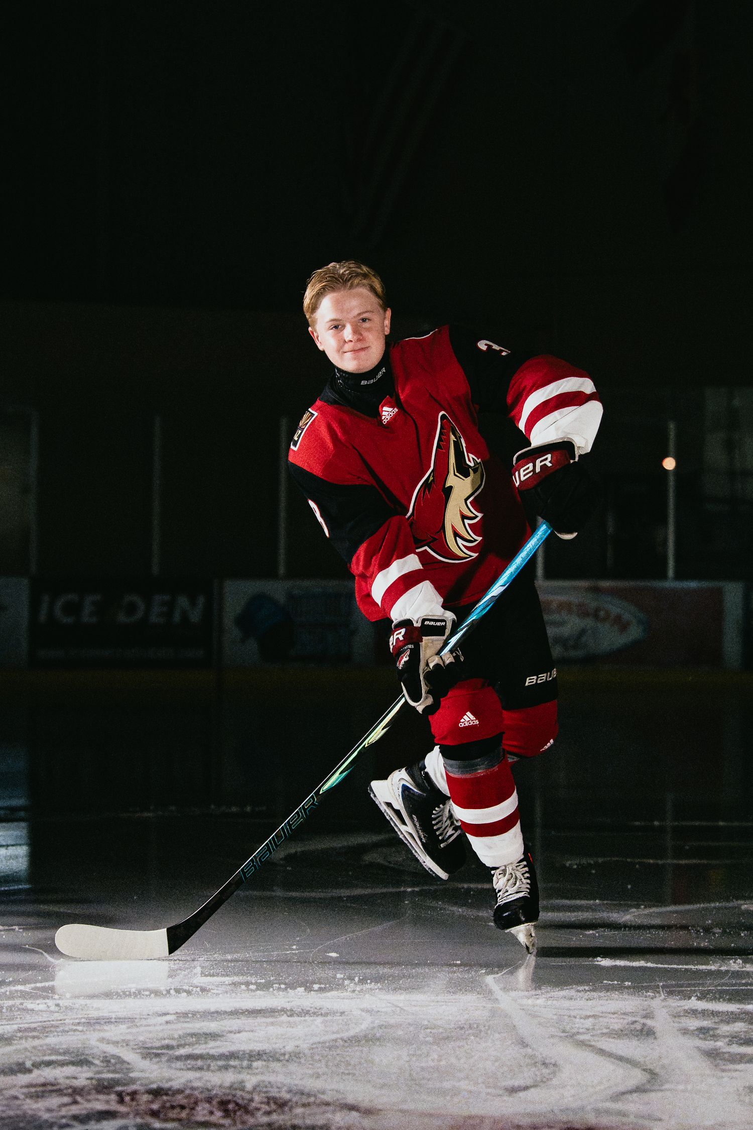 Hockey player in red Coyotes jersey poses on ice rink with stick at Ice Den arena.