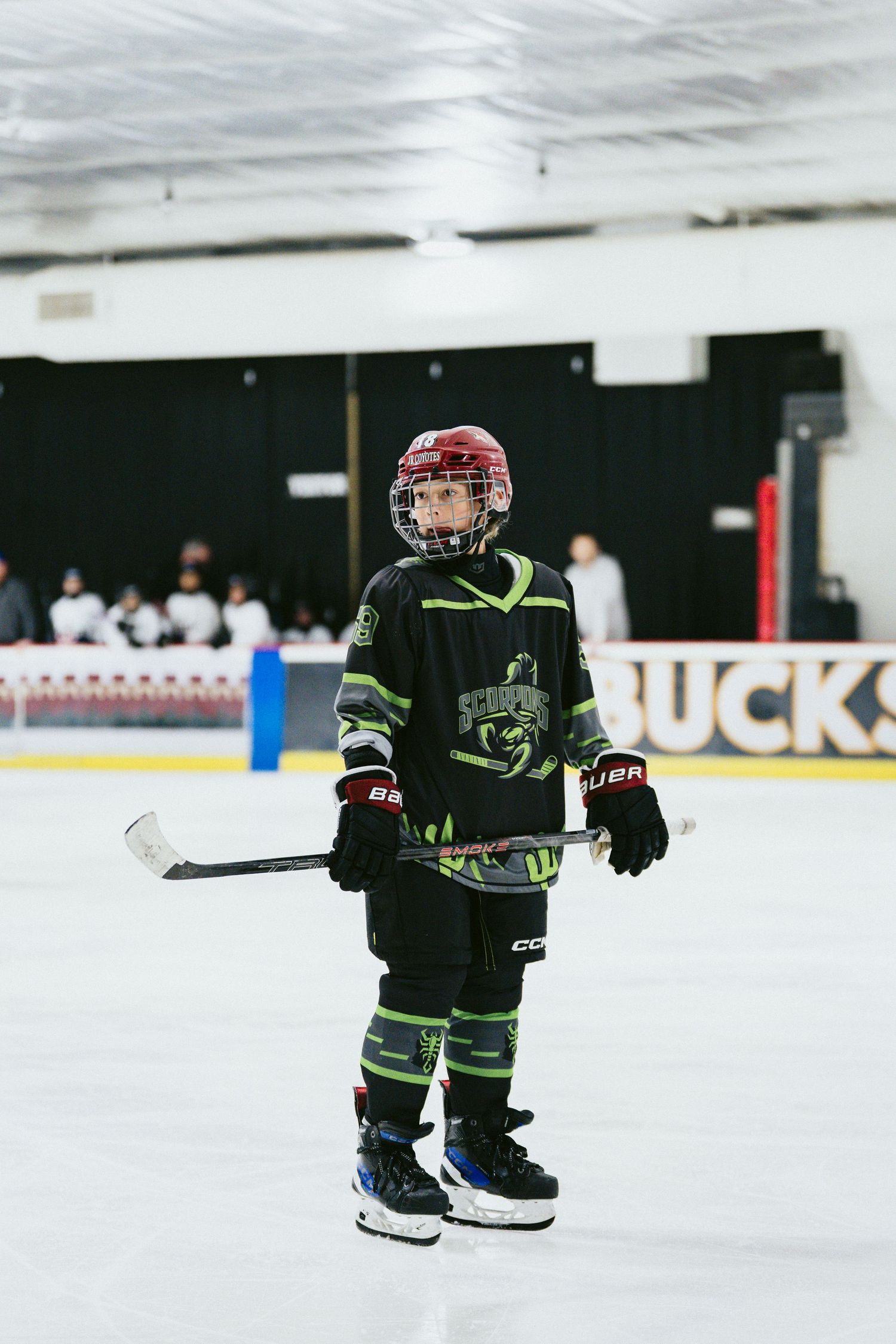 Young hockey player in black Bauer gear and red helmet standing on ice holding stick.