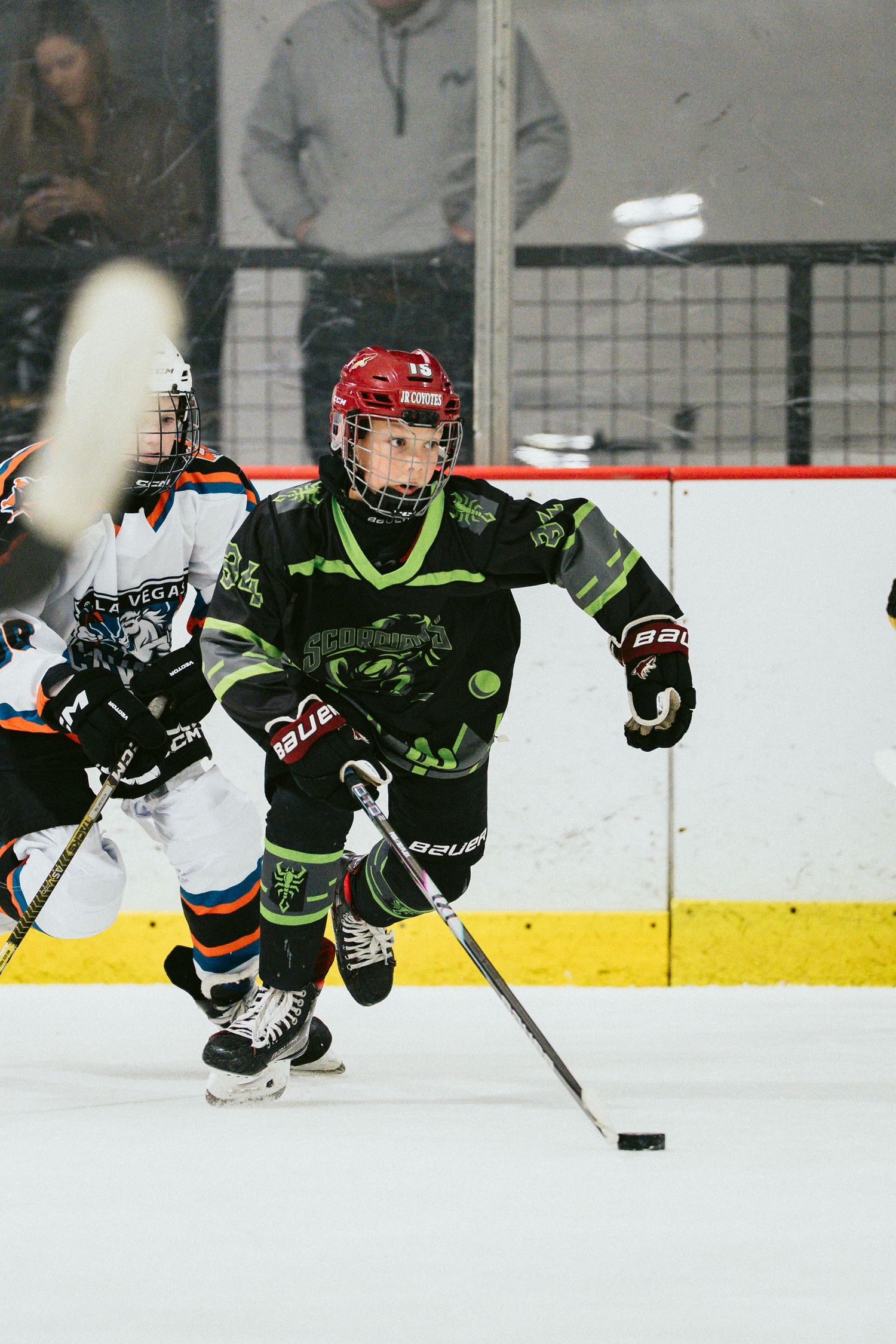 Youth hockey player in black green jersey controls puck while skating past opponent during indoor ice hockey game.
