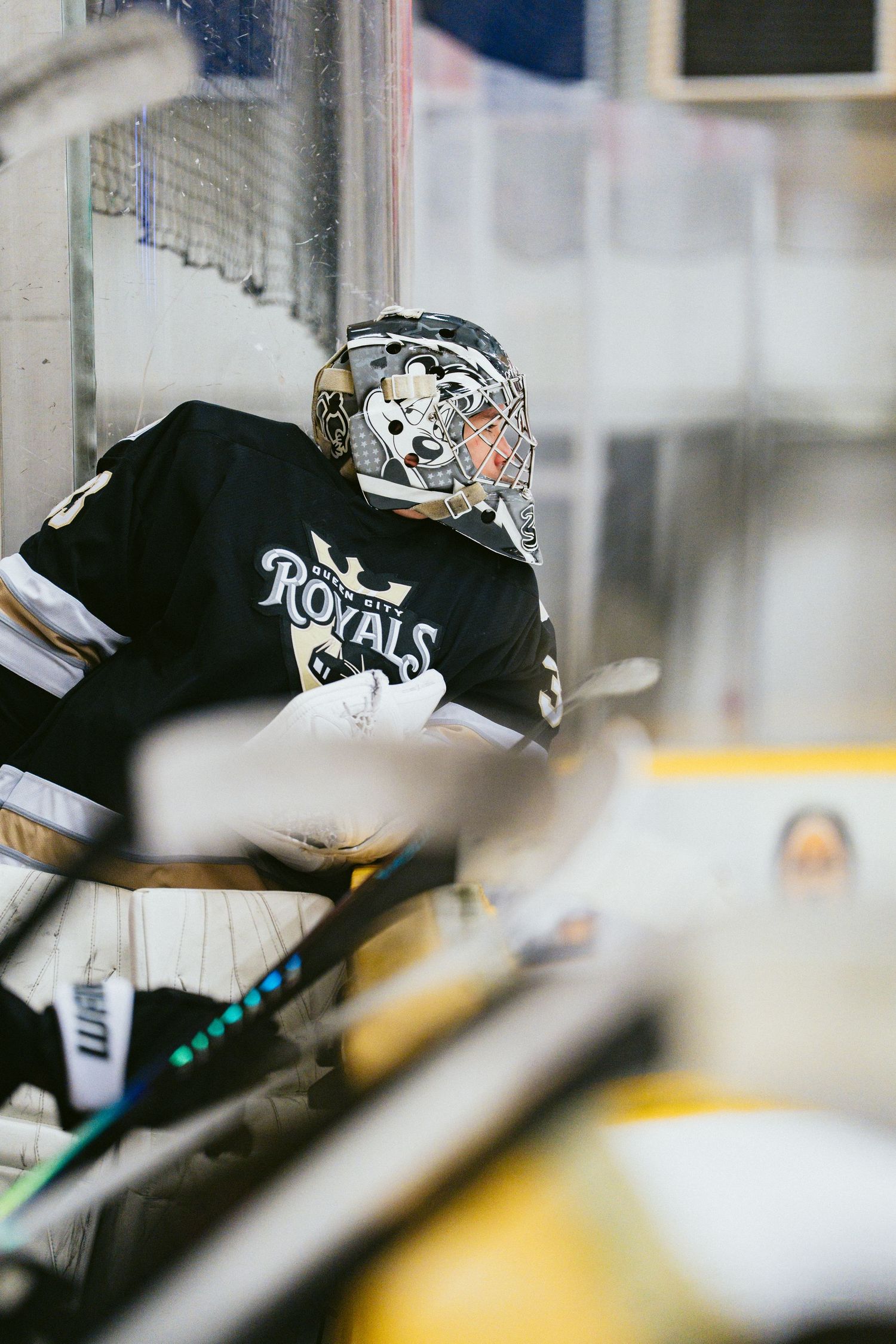 A hockey goalie in a black Royals jersey and decorated mask sits in the penalty box during a game.