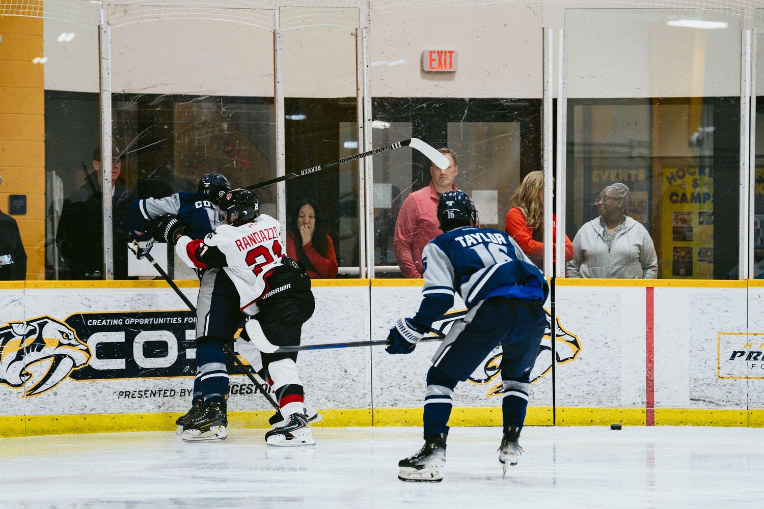Hockey players compete on an indoor ice rink during a game, with spectators visible behind the glass barrier.