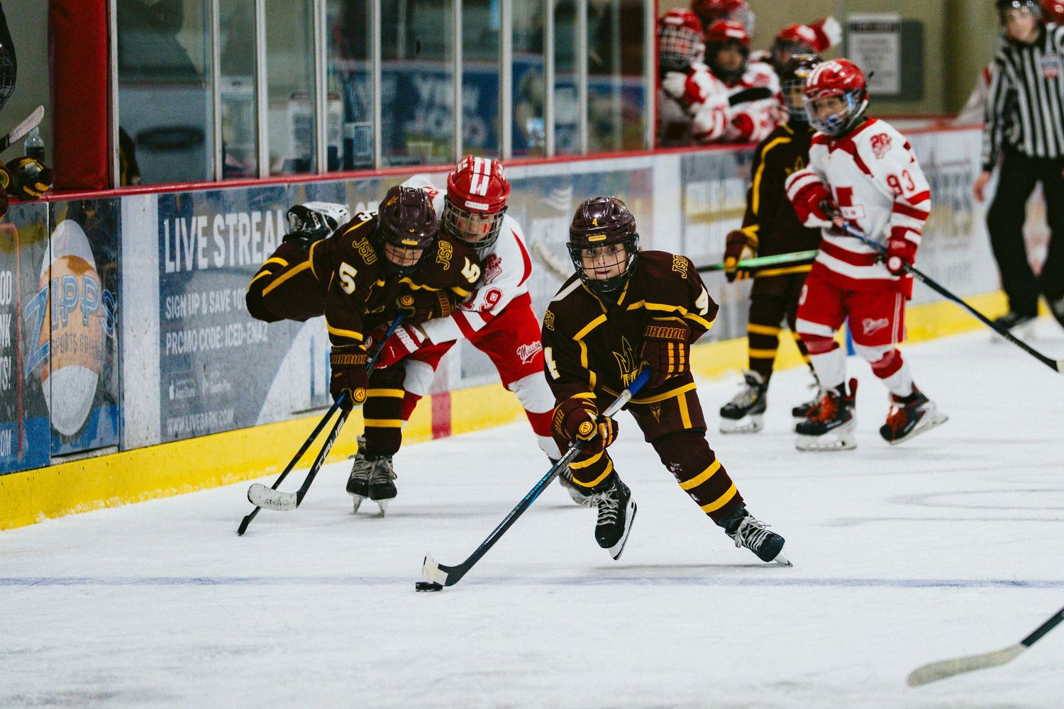 Youth hockey players in brown and red jerseys battle for the puck along the boards during a competitive game.