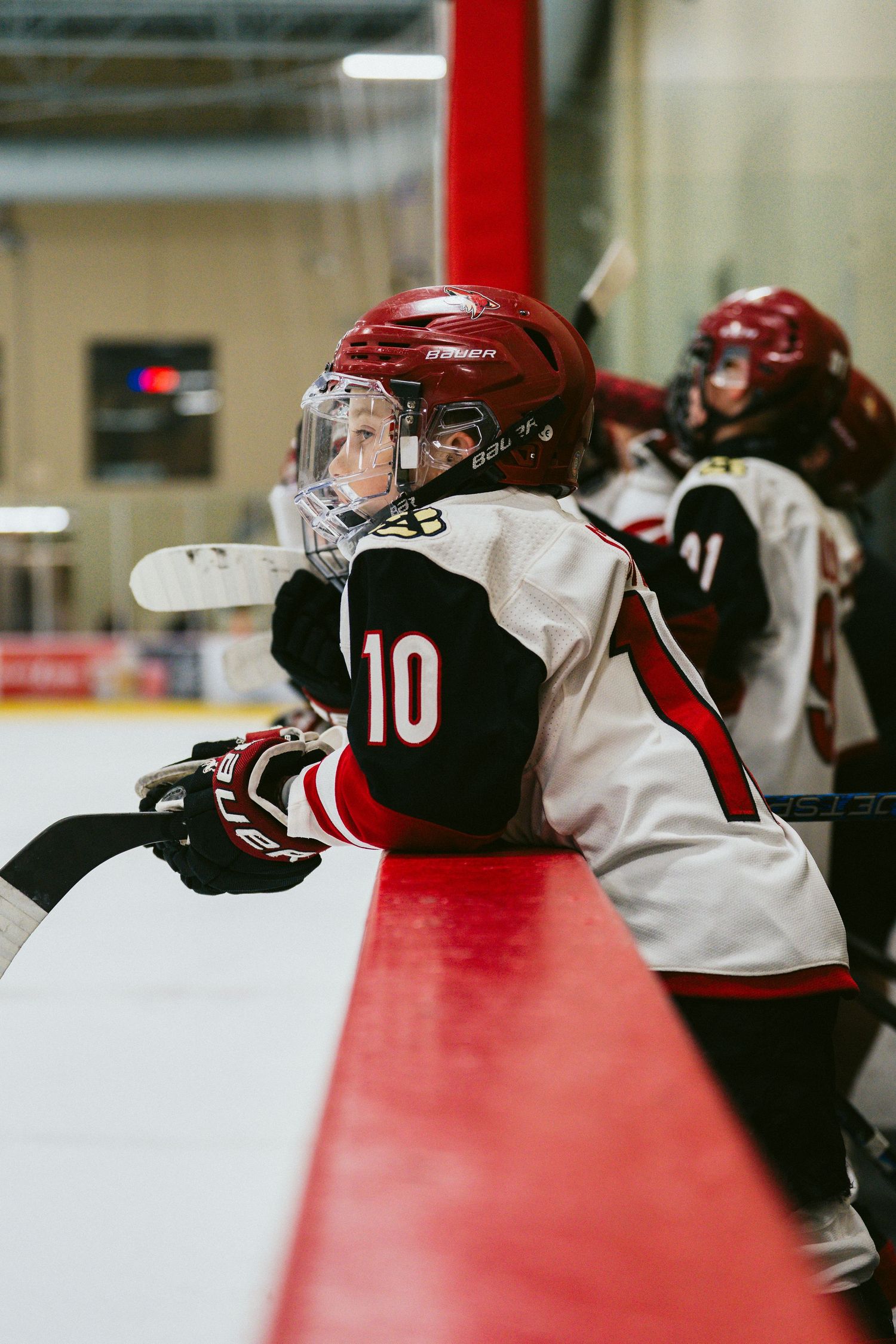 Youth hockey player in red helmet and white jersey watches game from player bench.
