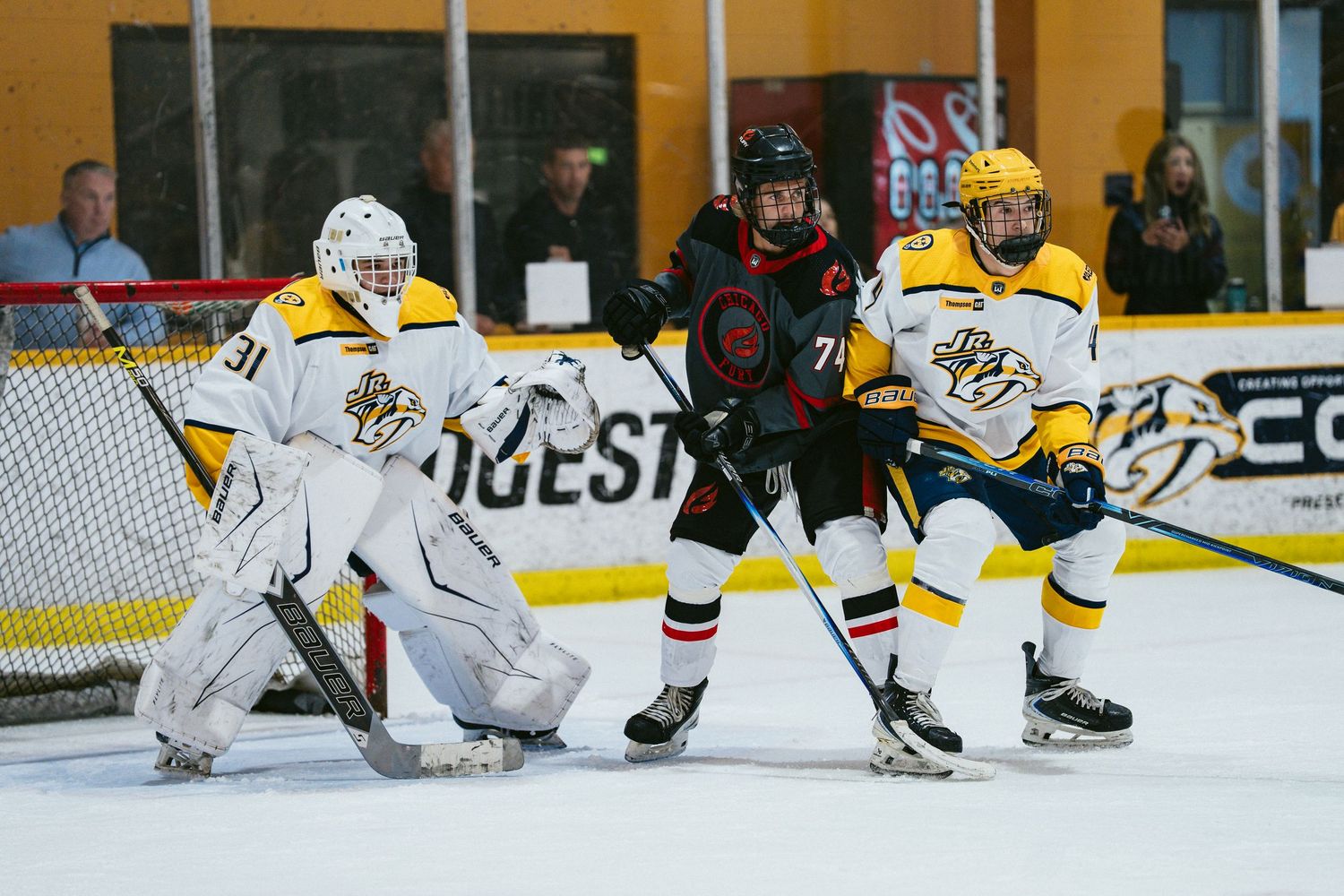 A goalie and defenseman in gold uniforms defend the net against an opposing player in black during a game.