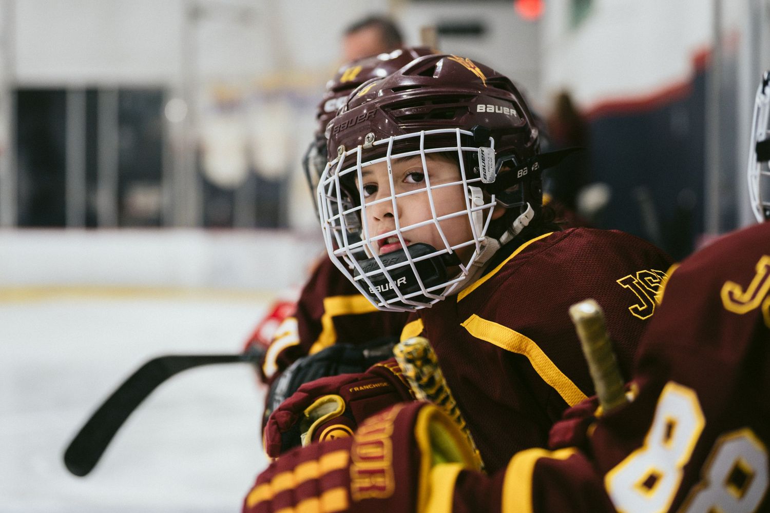 Young youth hockey player in maroon and gold uniform with cage helmet sitting on bench during ice hockey game.