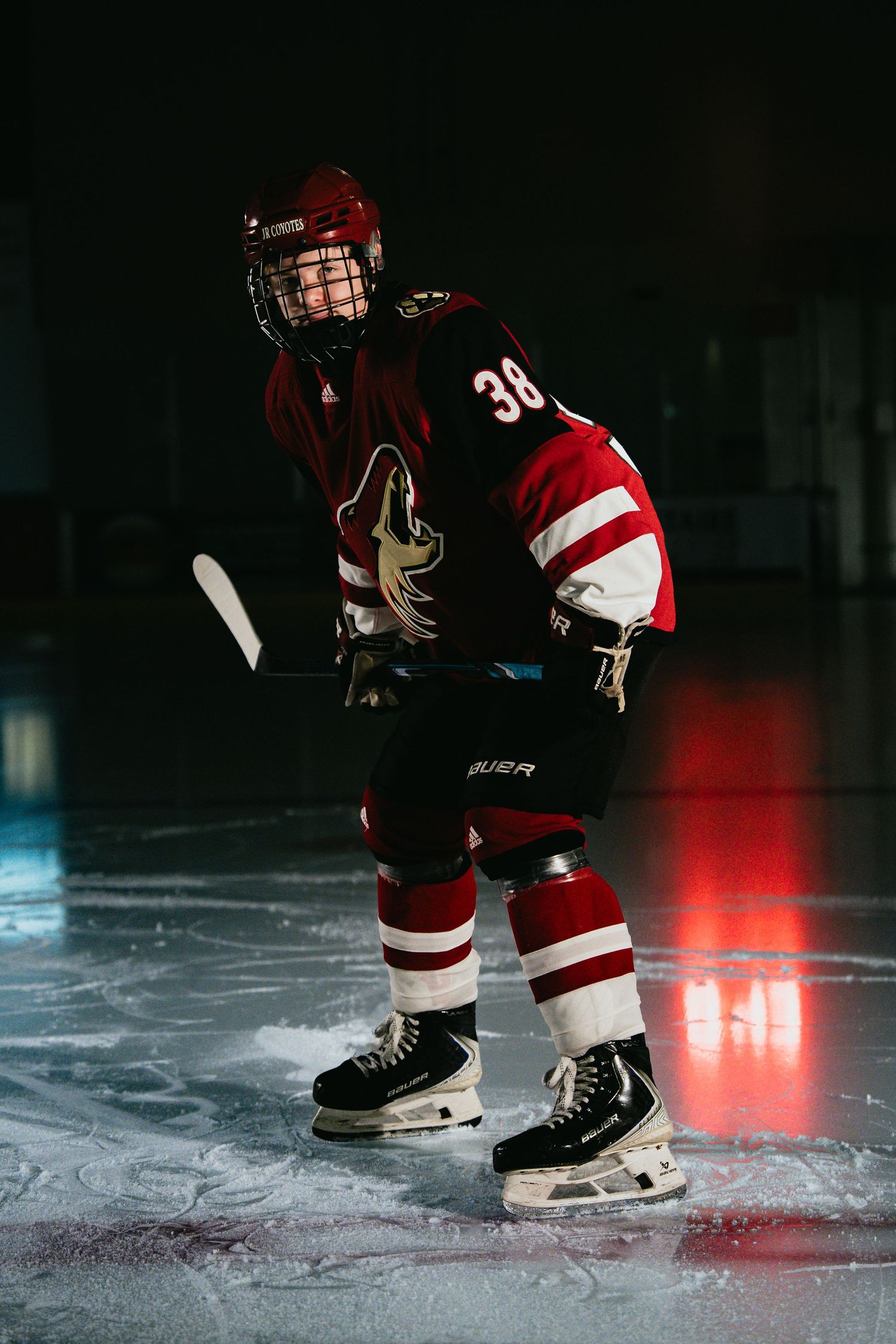 Young hockey player in red jersey number 38 stands on ice rink wearing Bauer skates and helmet with face guard.