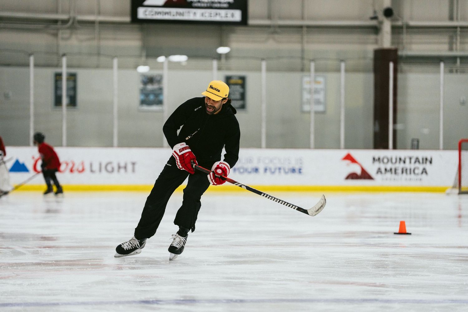 A hockey coach in black demonstrates skating technique around an orange cone on an indoor ice rink.