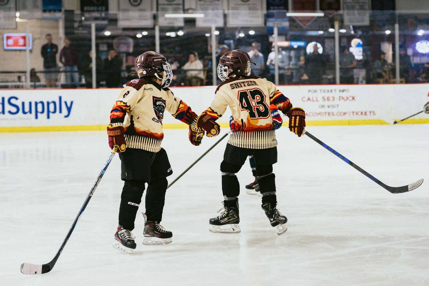 Two young hockey players in matching uniforms celebrate together on the ice during a youth game.