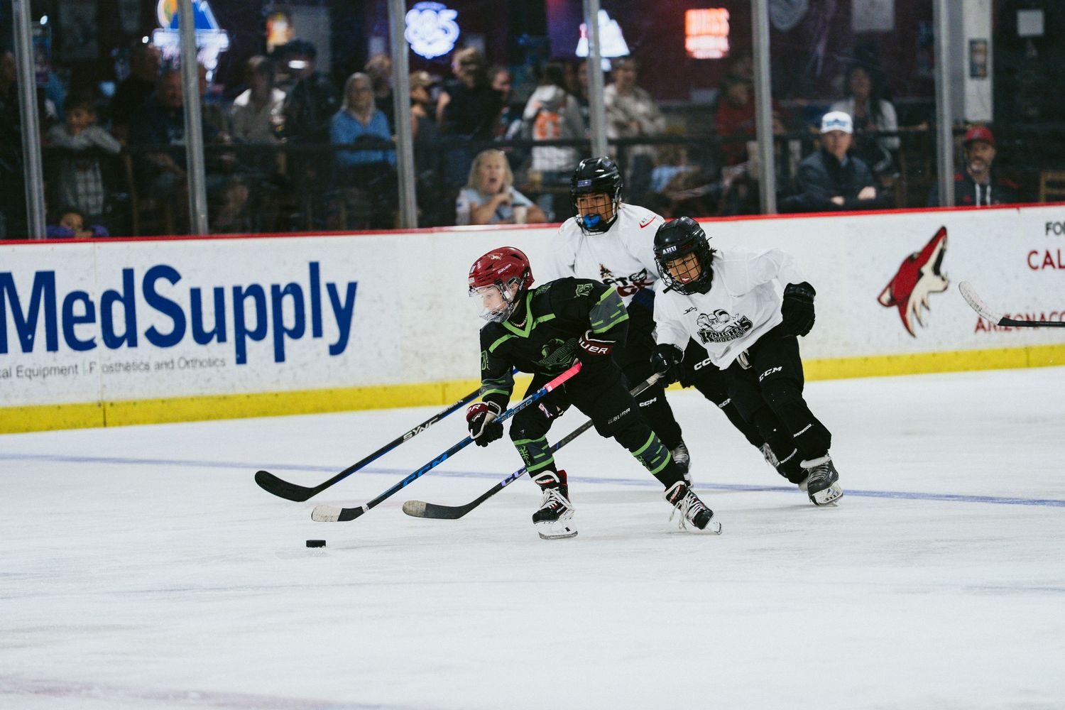 Youth hockey players in black uniforms compete for puck on indoor ice rink with MedSupply advertisement visible.