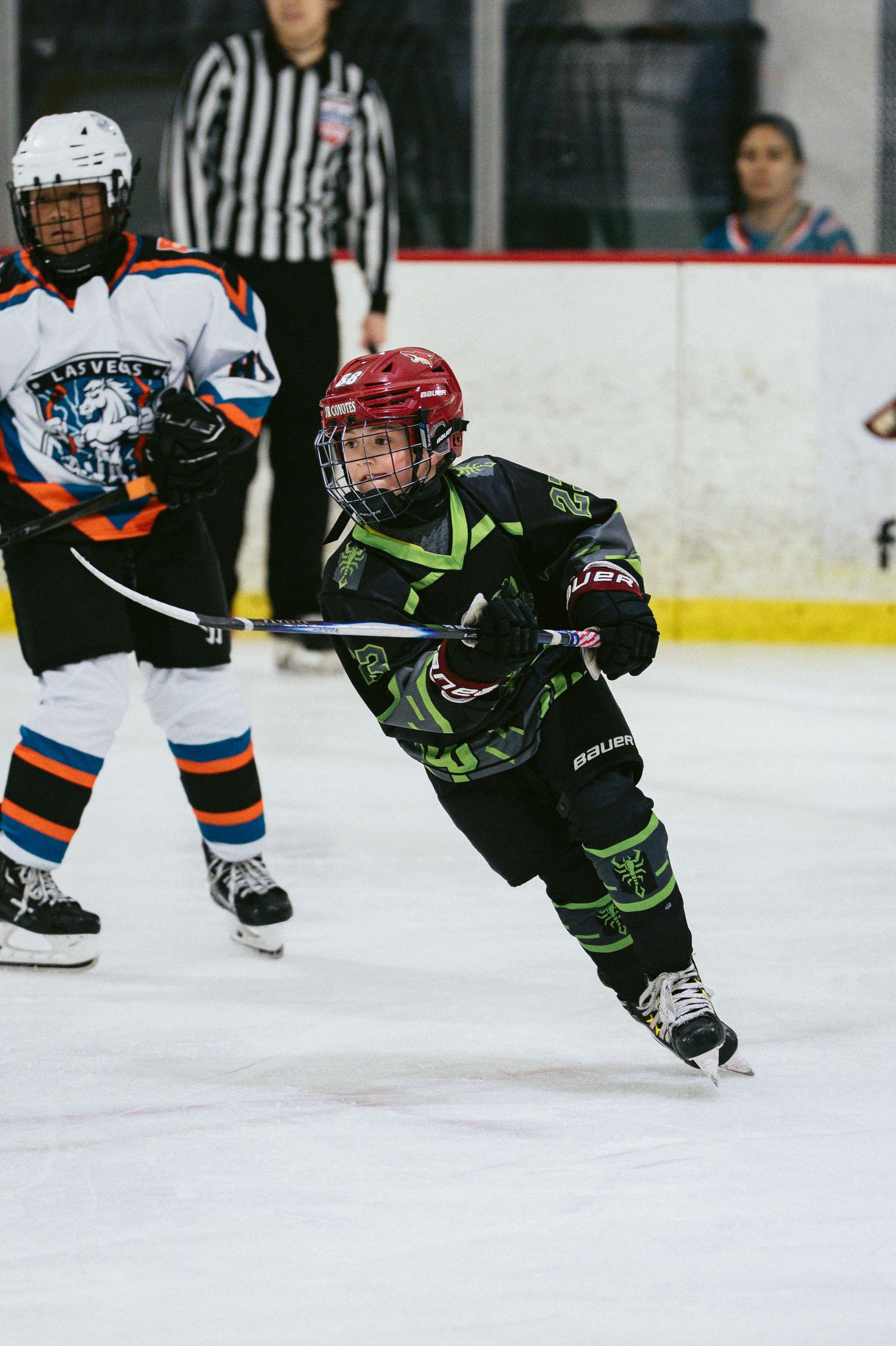 Young ice hockey player in black and green uniform skating during a youth hockey game with referee in background.