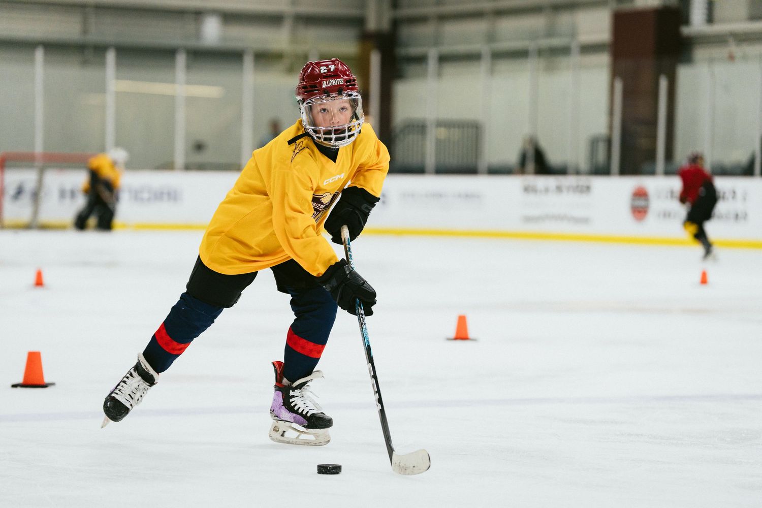 Youth hockey player in yellow jersey skating with puck through orange cones during indoor drill.