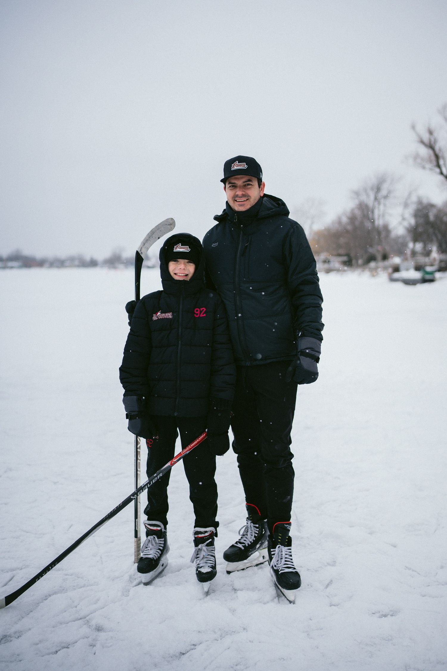 Two people in winter coats stand together on snowy ground holding hockey sticks.