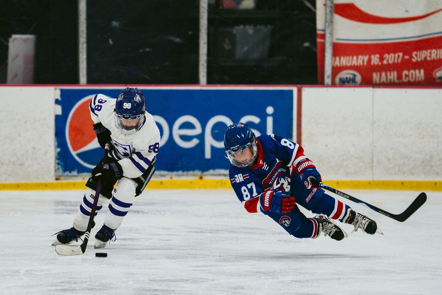 Youth hockey player in white and blue uniform skating on ice during a game at an indoor arena.