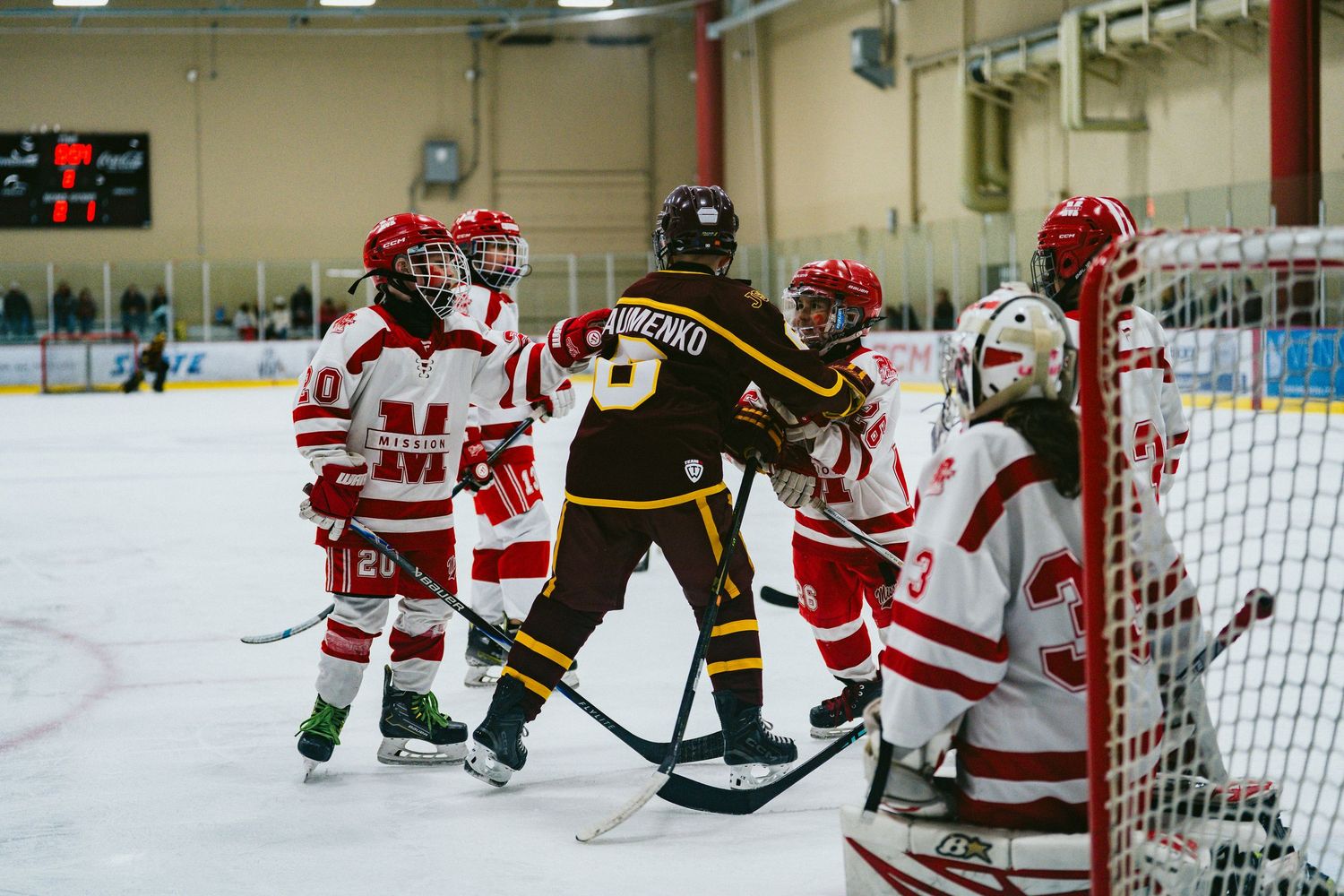 Youth hockey match between teams in red and black uniforms with players gathered around the goal.