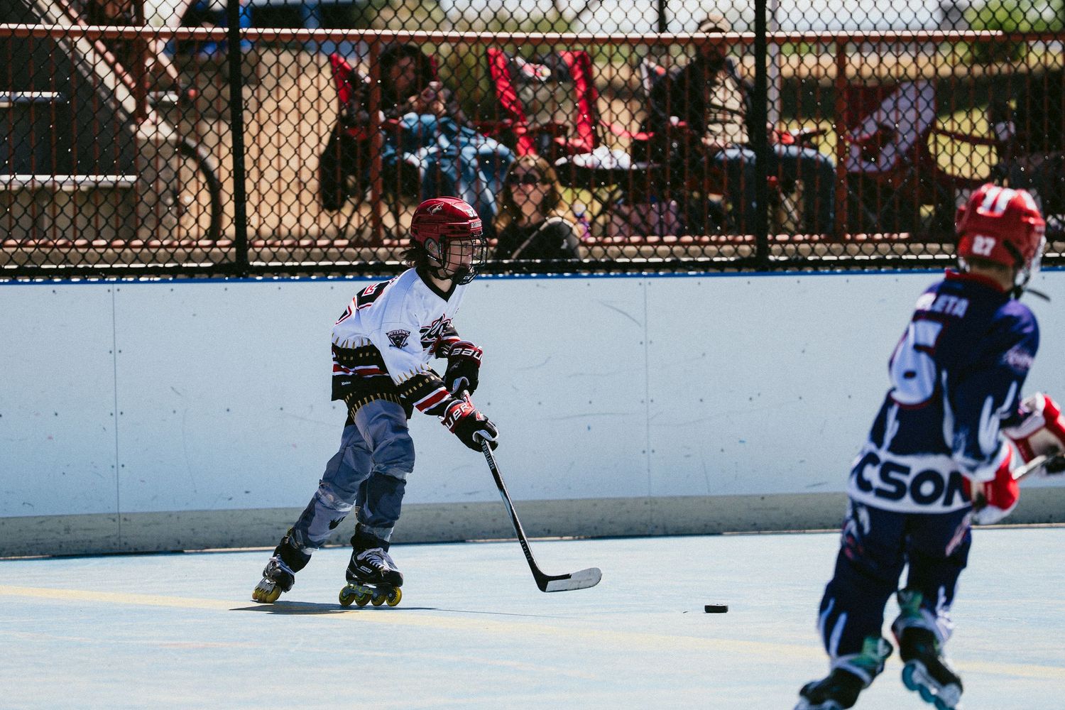 Young child in red helmet plays roller hockey on outdoor rink, holding stick near puck.
