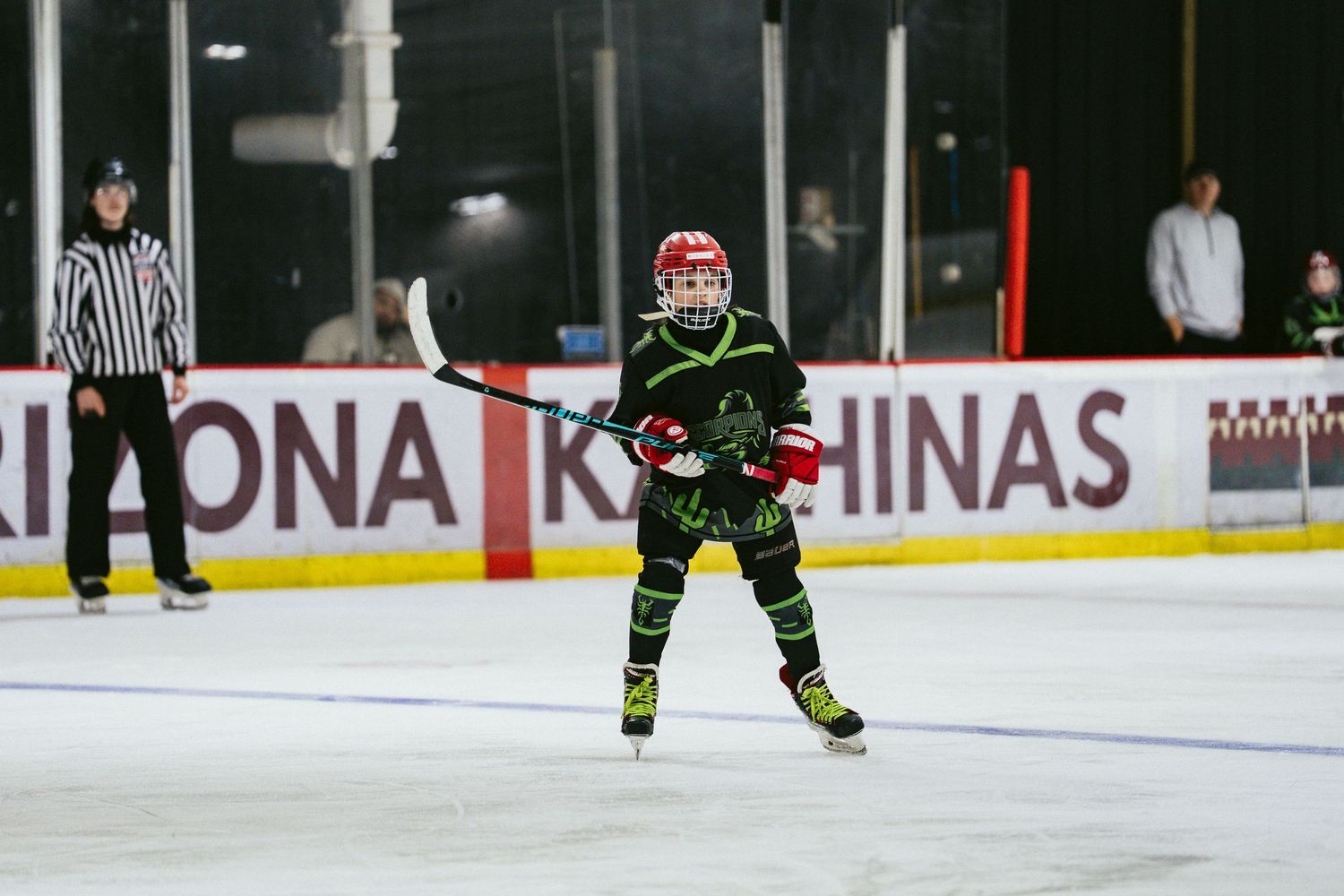 Young hockey player in black green uniform and red helmet skates on ice with referee visible in background.