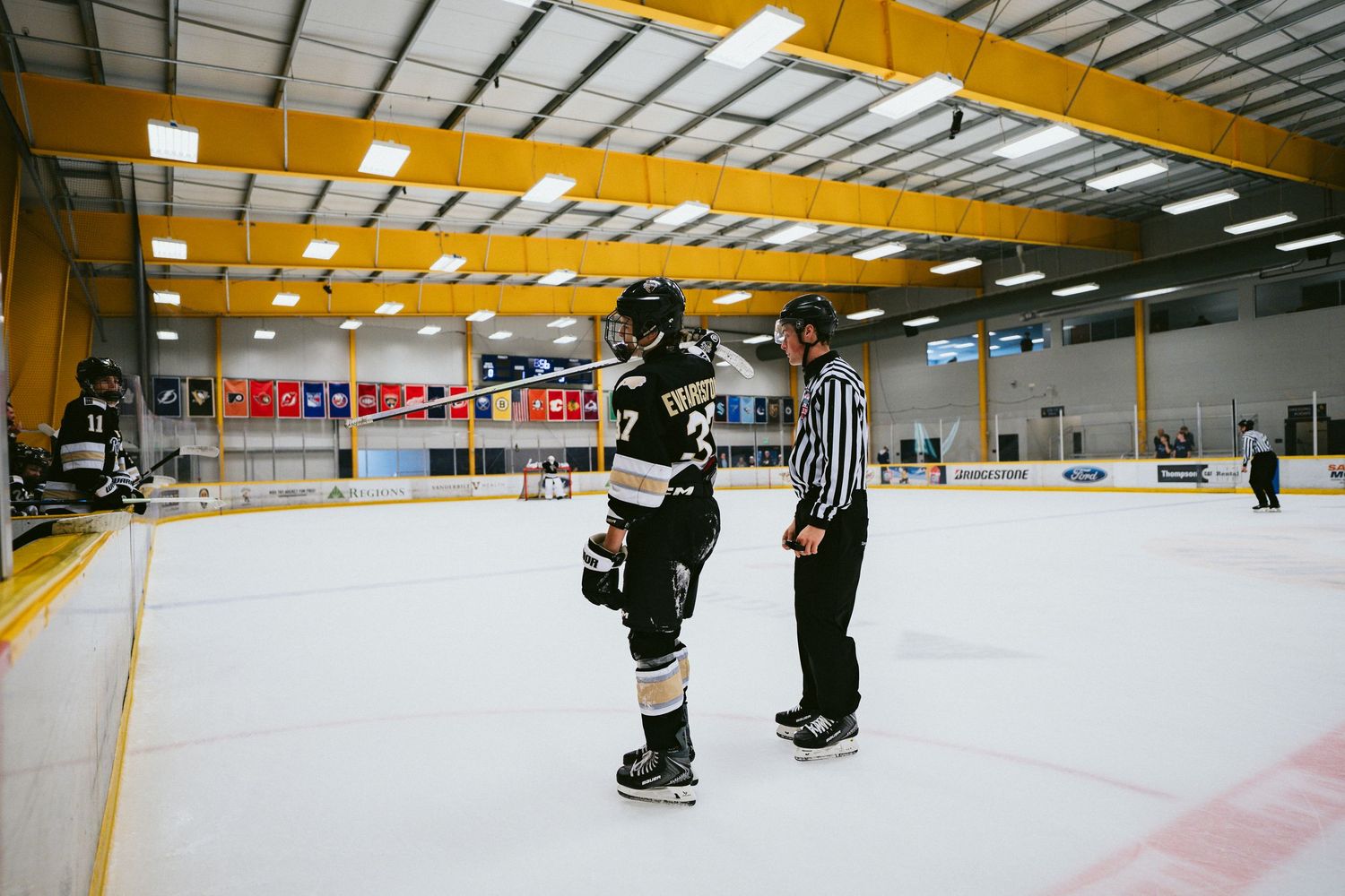 Hockey player in black uniform consulting with a referee on an indoor ice rink during a game.