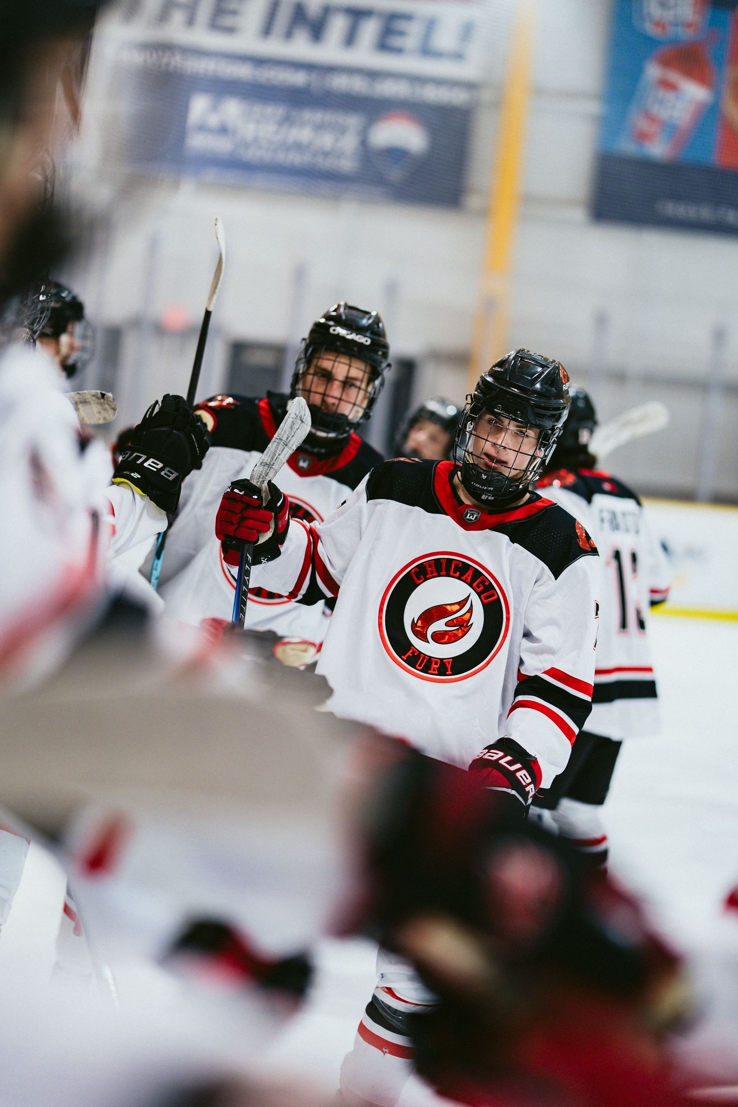 Youth ice hockey players in white uniforms celebrate on the rink, holding sticks up in team huddle.