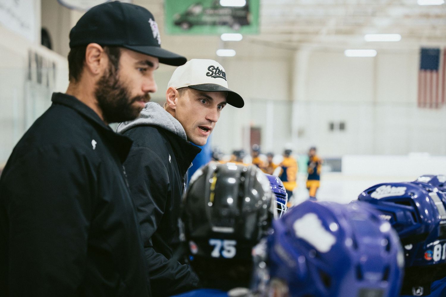 Two hockey coaches in black jackets discuss strategy on the bench during youth ice hockey game.