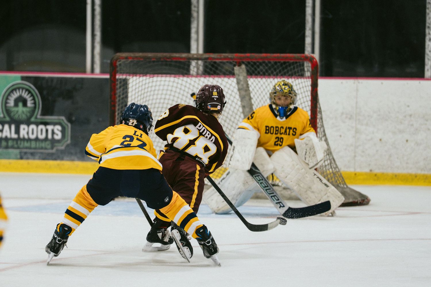 Hockey player in maroon jersey attacks the net against a Bobcats goalie during an indoor ice hockey game.