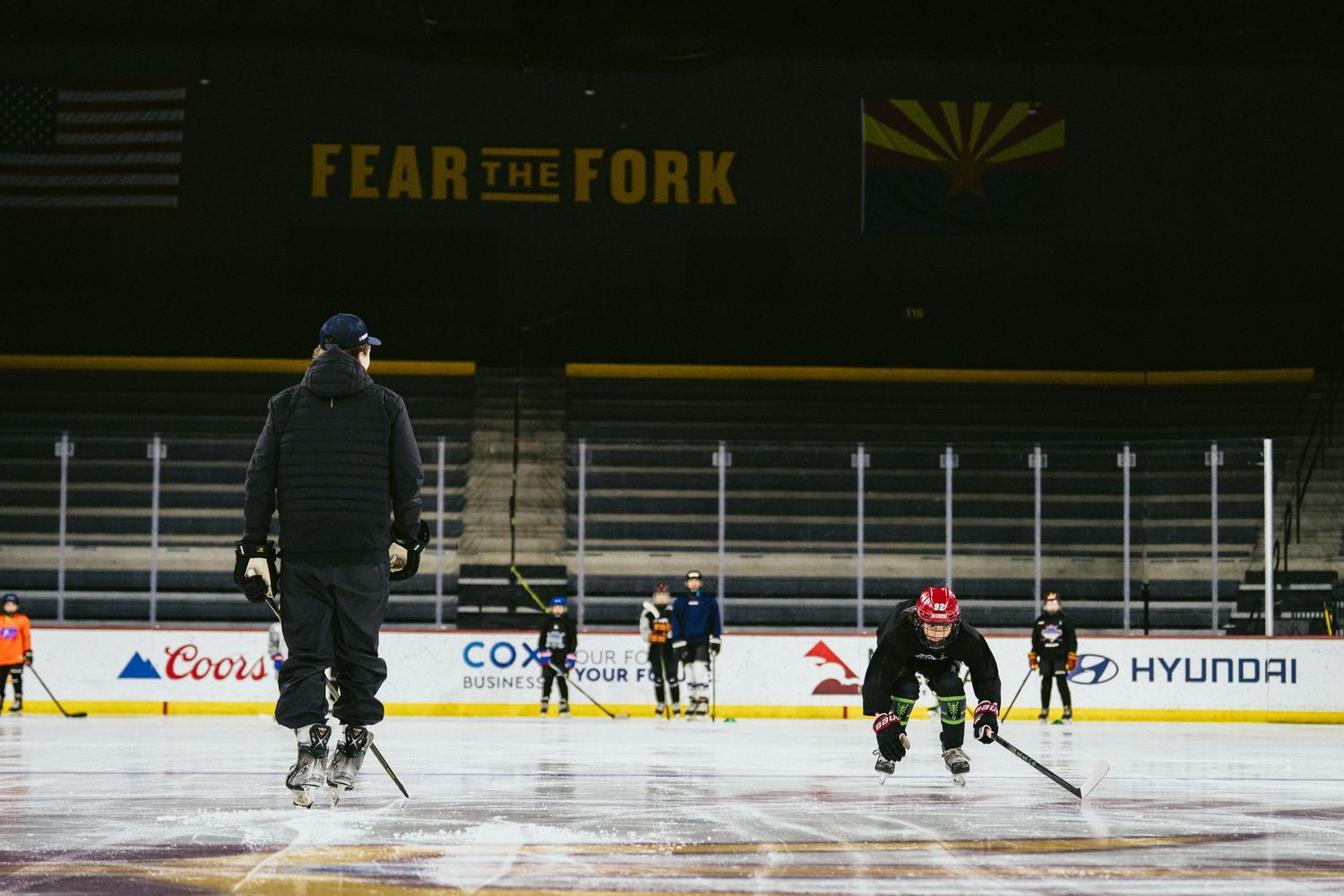 Youth hockey players practice skating drills on an NHL rink beneath a Fear the Fork banner in Arizona.