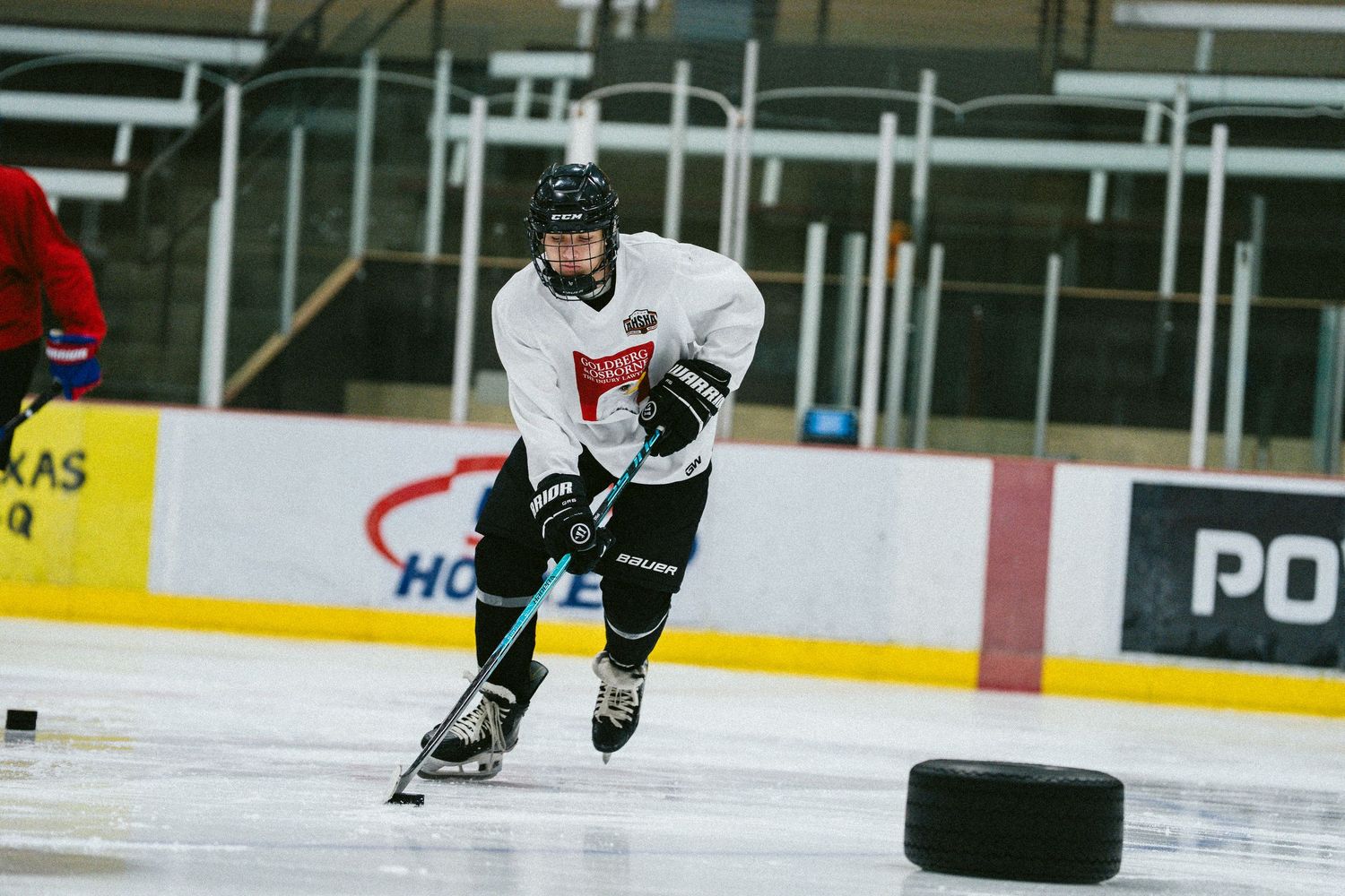 Hockey player skating with puck during indoor ice rink practice session wearing white jersey.