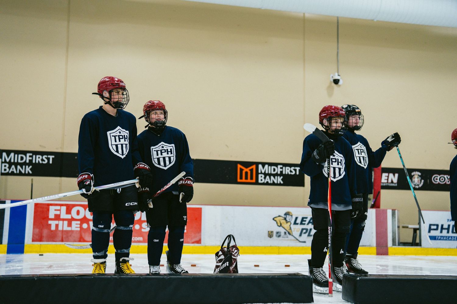 Four youth hockey players in black uniforms and red helmets stand on the ice rink bench during a game.