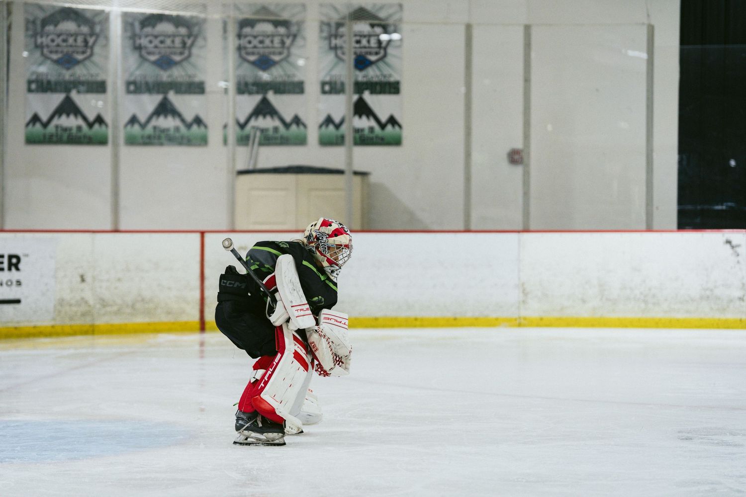 Hockey goalie in crouch position on indoor ice rink with mountain banners in background.