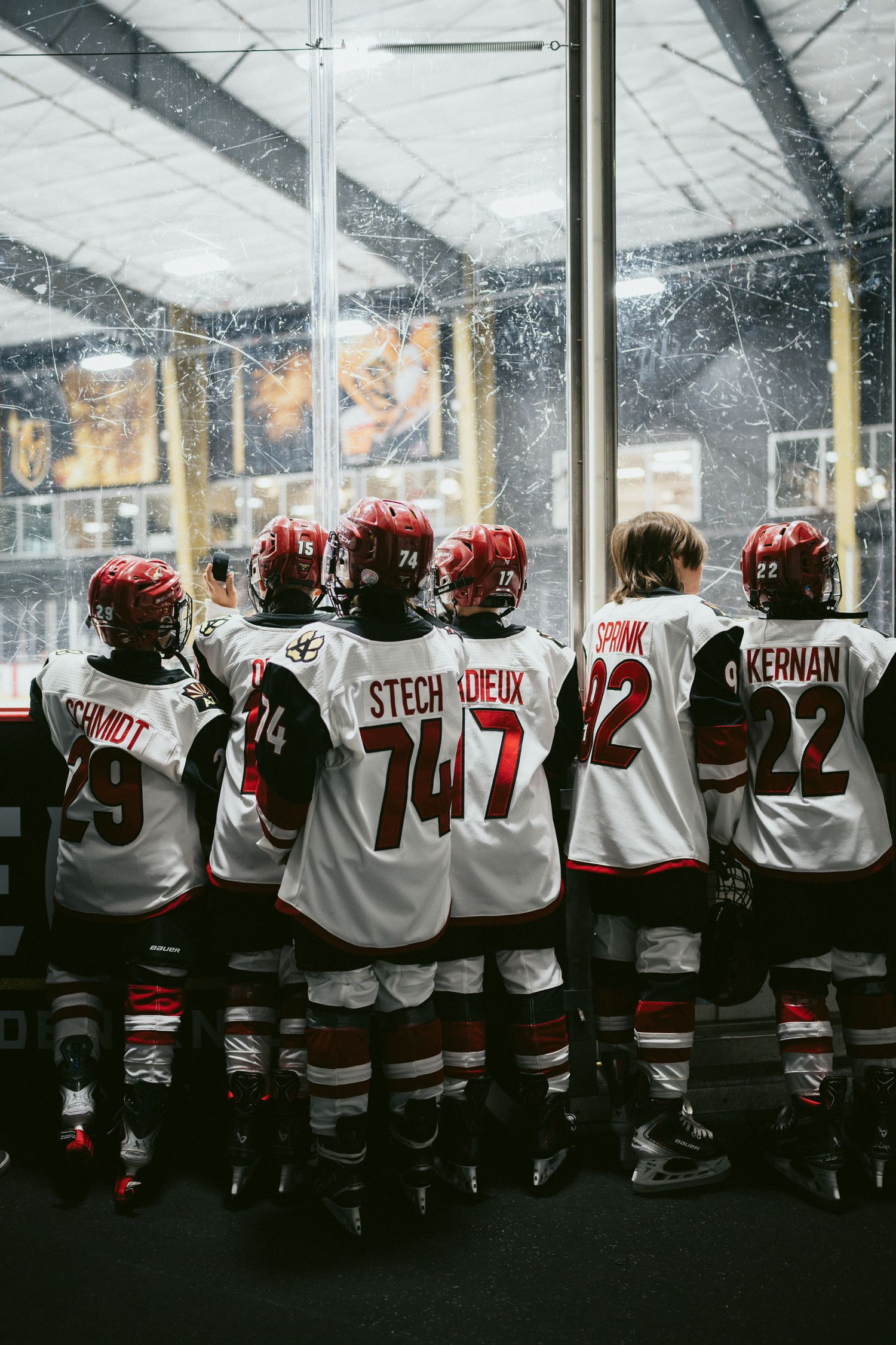Hockey players in white jerseys with red helmets watching through glass at ice rink.