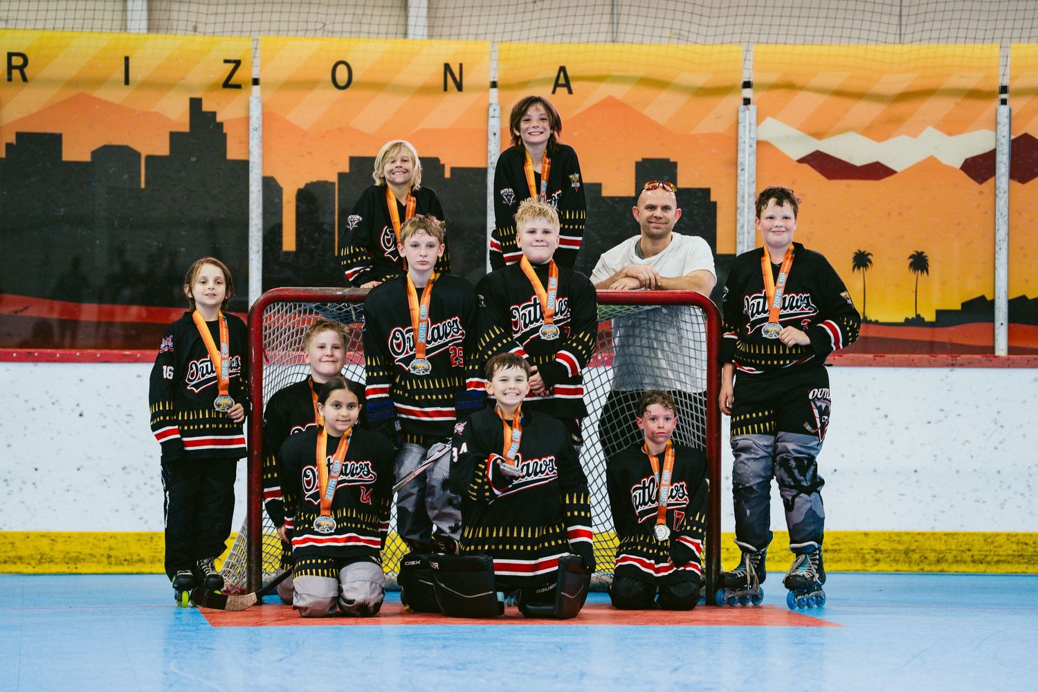 Youth roller hockey team in black jerseys poses with medals and coach in front of a goal at an Arizona rink.