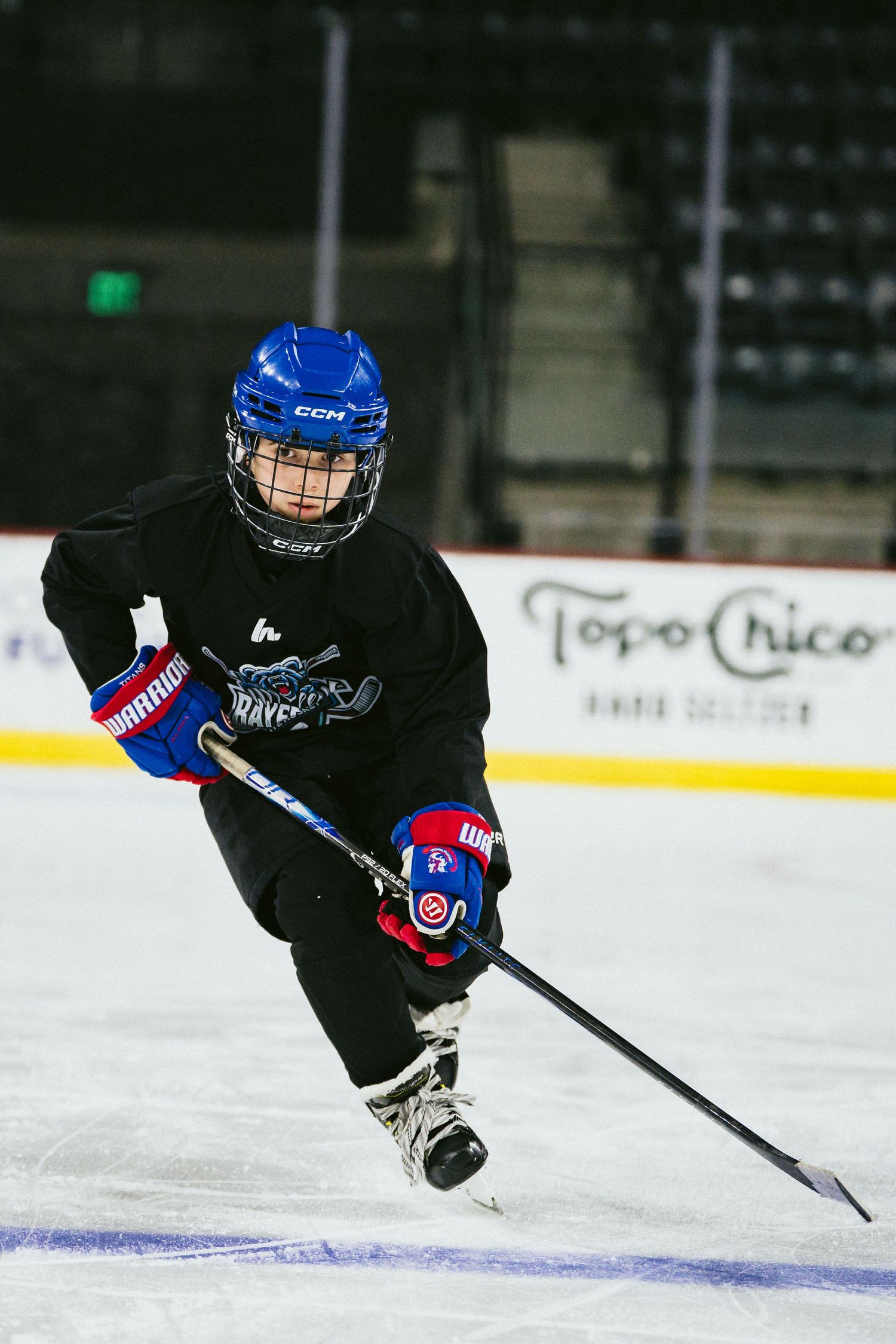 Young hockey player in blue CCM helmet and black jersey skating with stick on indoor ice rink.