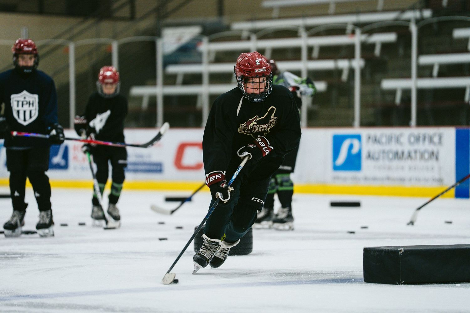 Young hockey player in black uniform executes low skating stride with puck on indoor rink.