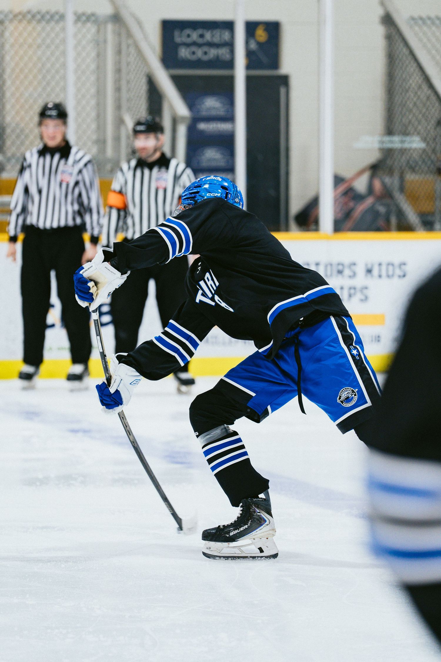 Ice hockey player in blue and black uniform takes a shot on ice with two referees visible in the background.