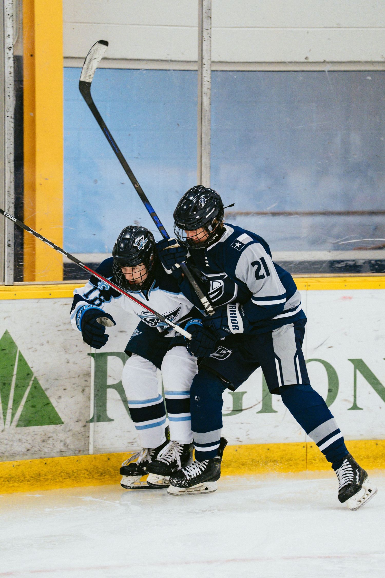 Two youth hockey players in white and navy jerseys battle along the boards during a competitive ice hockey game.