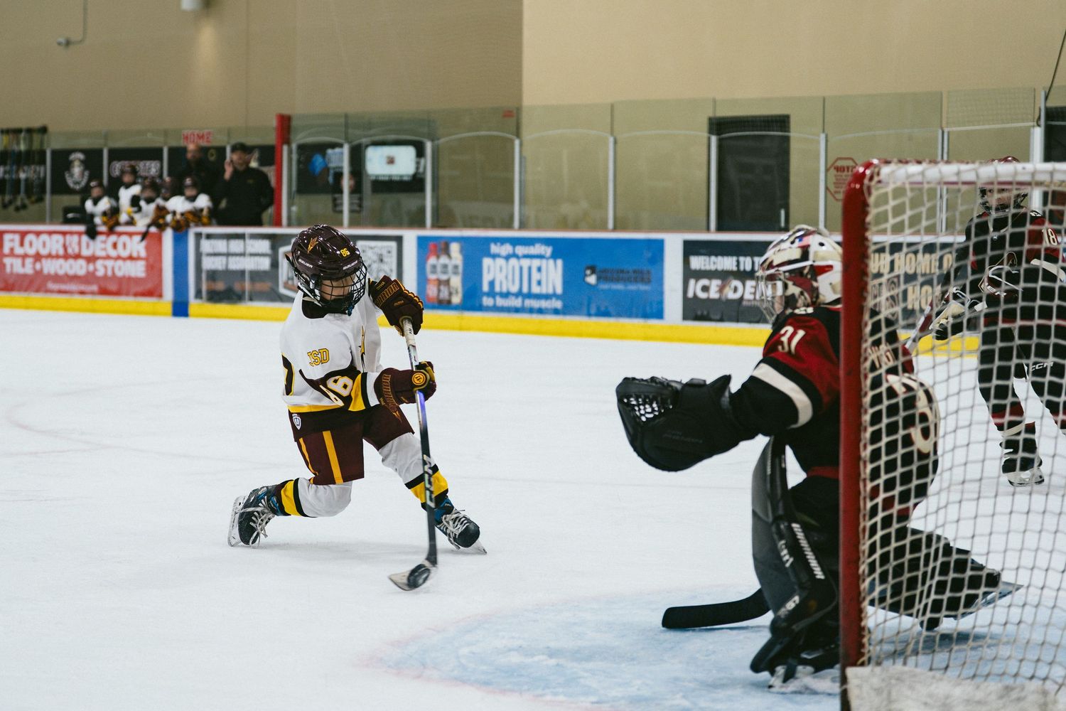 Youth hockey player in maroon and gold takes a shot on goal against a goalie in an indoor rink.