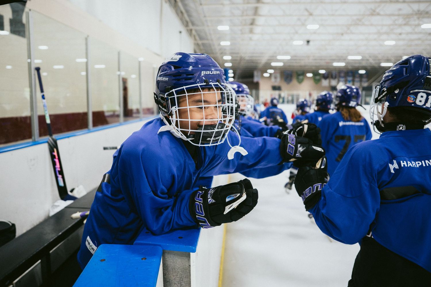Youth hockey players in blue uniforms fist bumping near the rink boards during a hockey practice session.