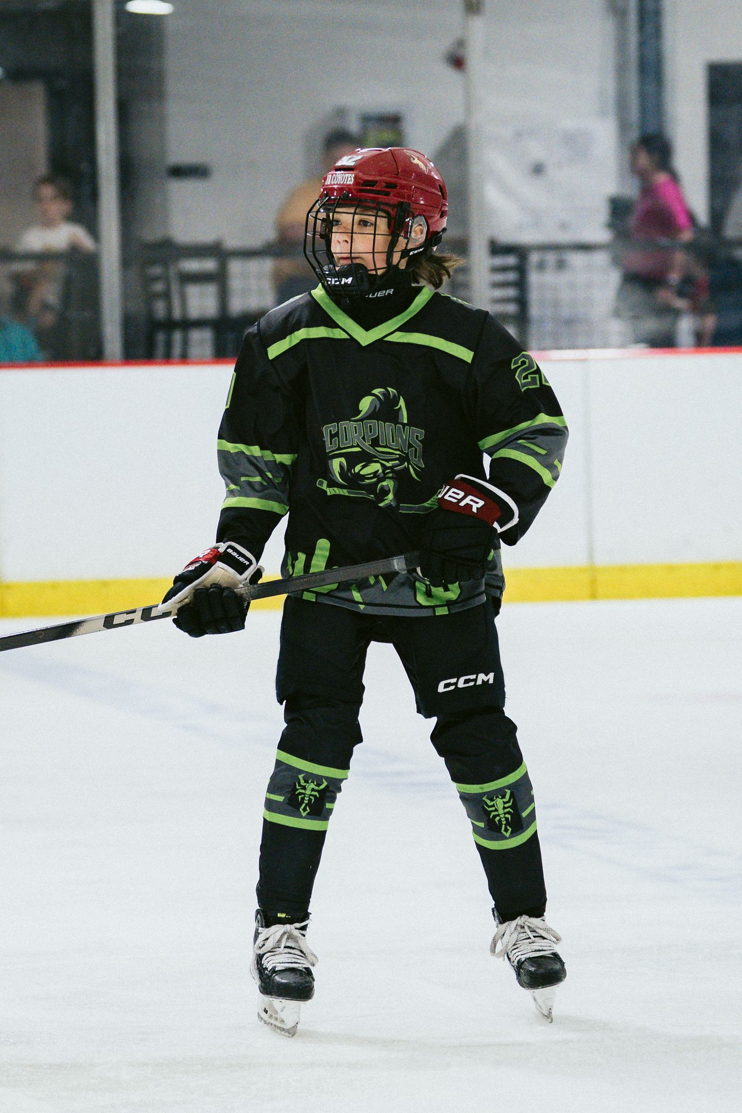 Young hockey player in black green Scorpions uniform and red helmet standing on ice rink.
