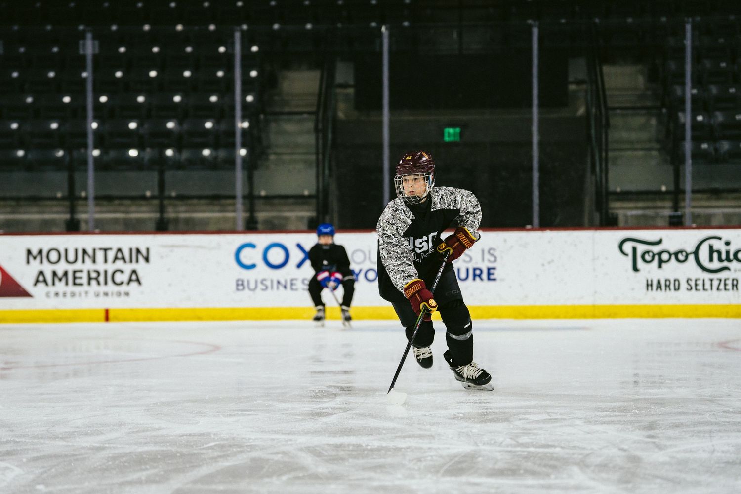Youth hockey player in camouflage jersey skating on indoor ice rink during practice with sponsor banners visible.