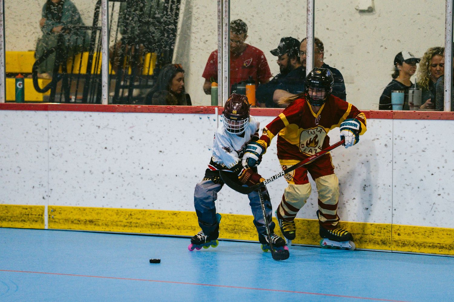 Two youth roller hockey players battle for the puck along the boards during an indoor inline hockey game.