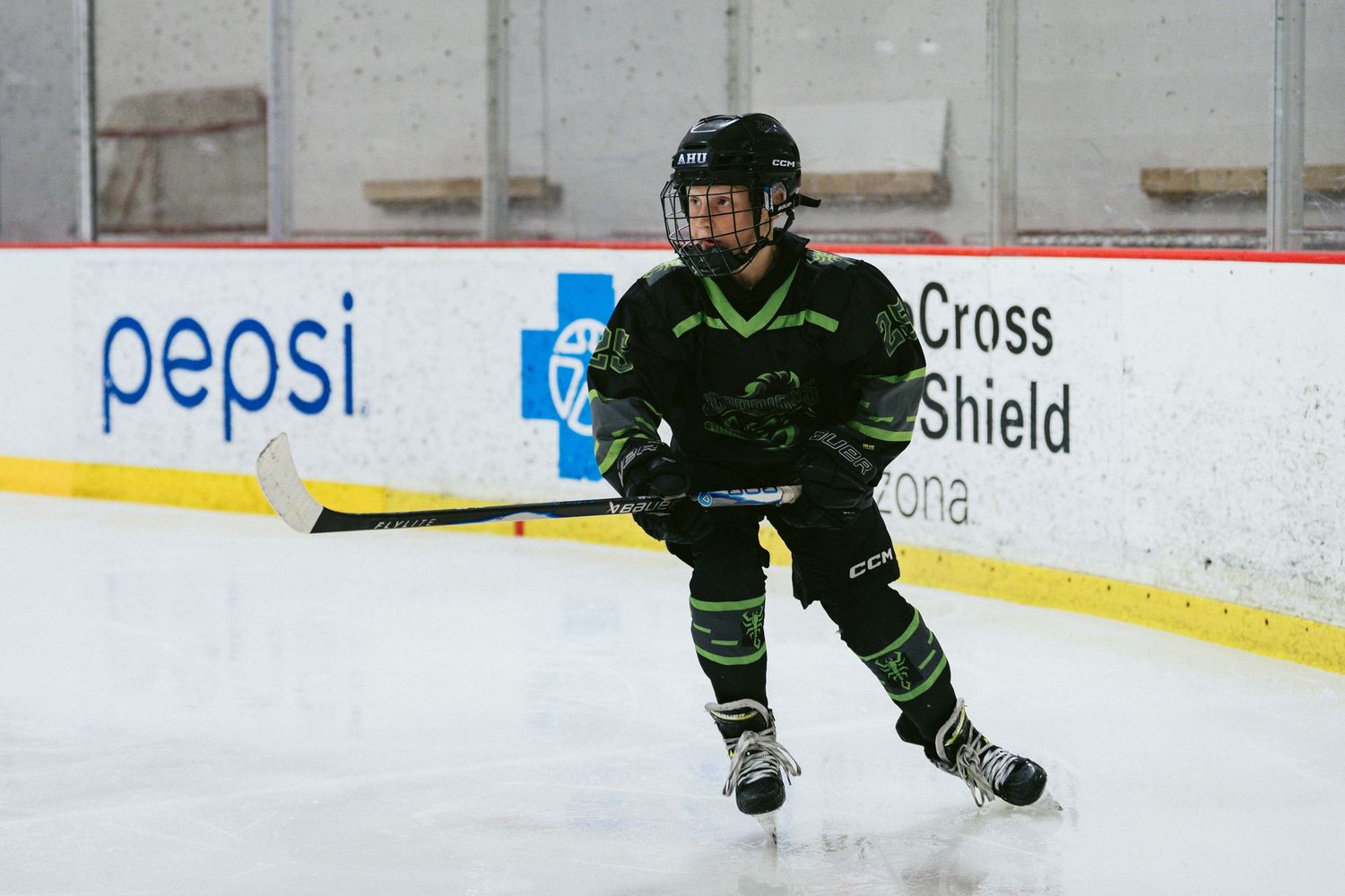 Young hockey player in black and green uniform practices skating skills on indoor ice rink with Pepsi ad visible.