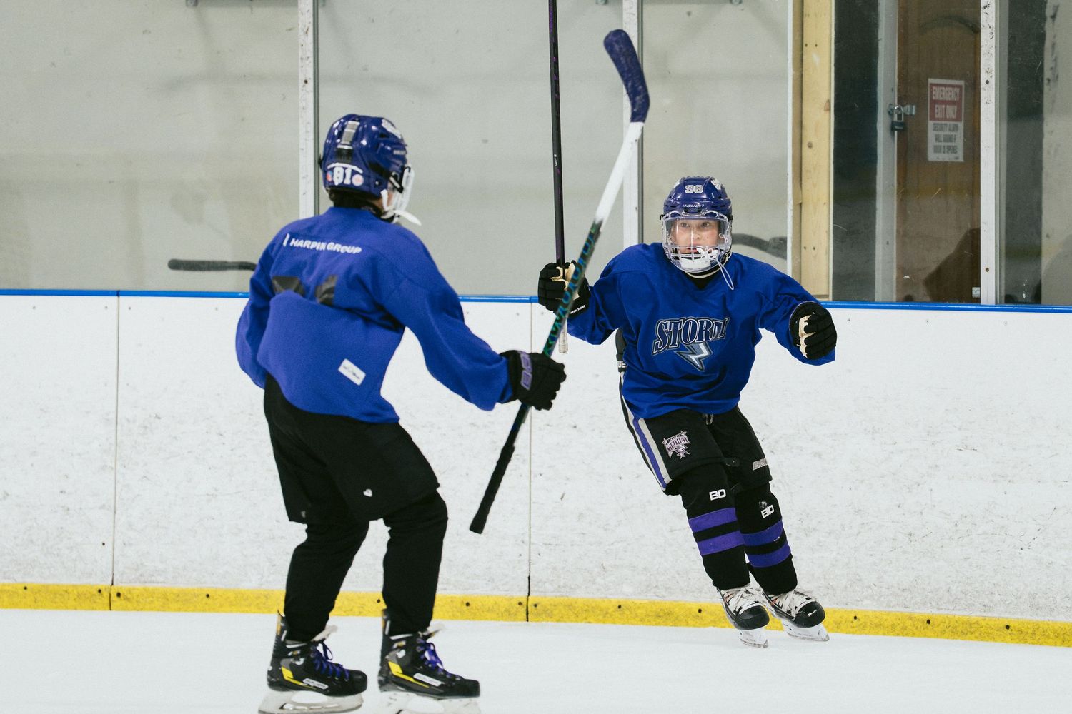 Two youth hockey players in blue uniforms practice passing drills on an indoor ice rink during team training.