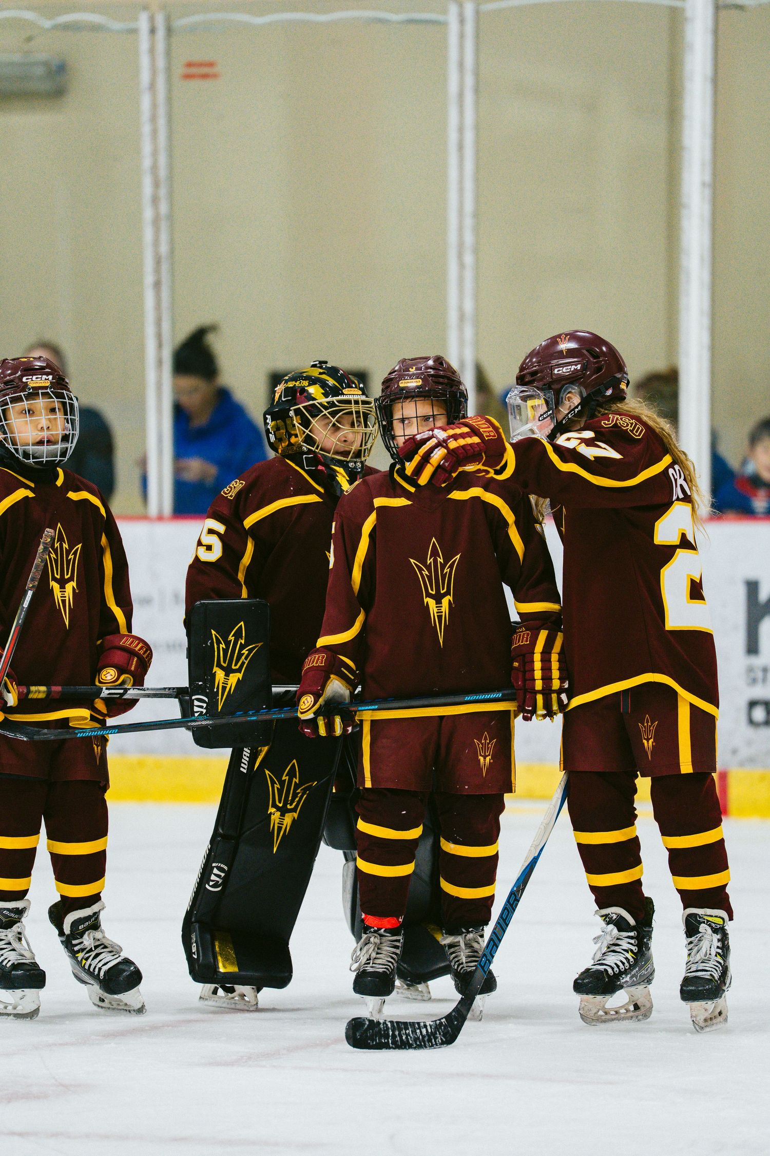 Hockey team players in maroon and gold uniforms with pitchfork logos gather on the ice during a game.