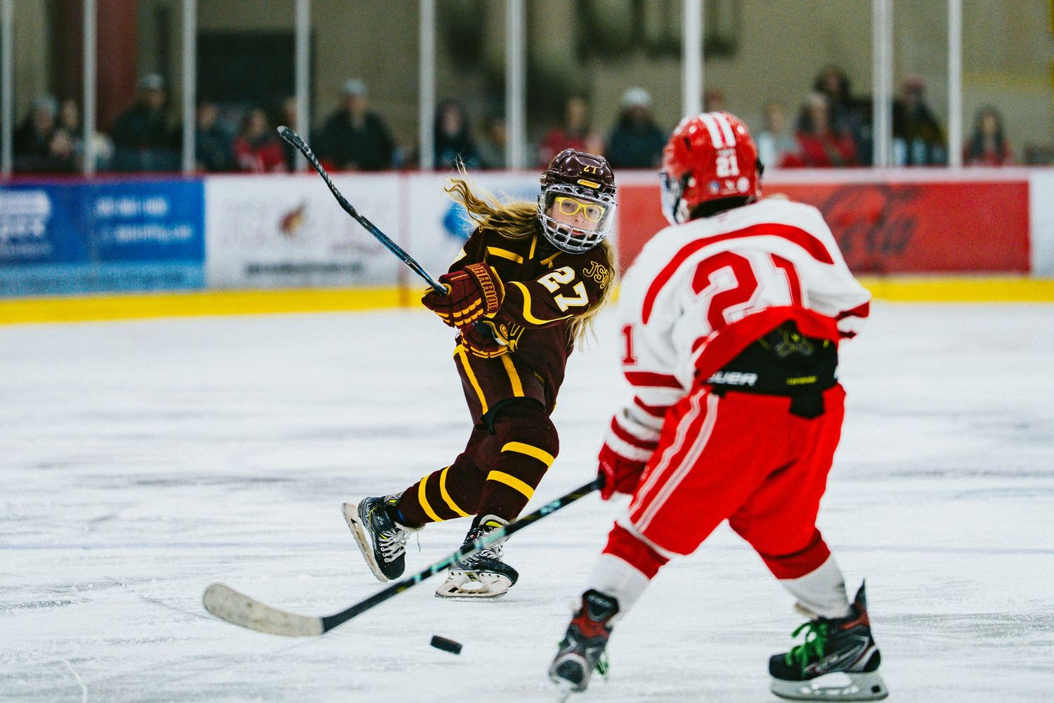 Hockey players in black and red uniforms chase after loose puck during competitive game action.