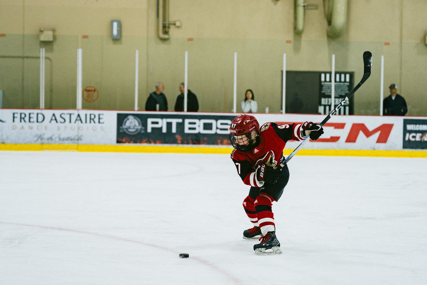 Hockey players skate on ice during a game with advertising boards visible along the rink walls.