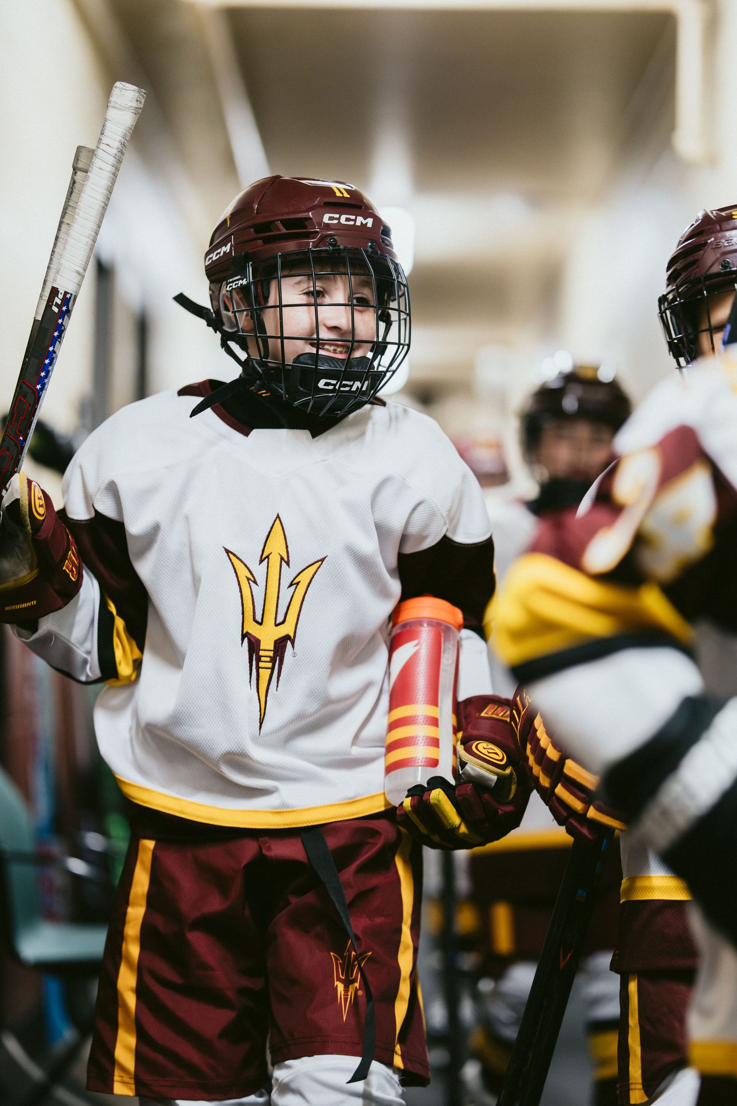 Smiling young hockey player in Arizona State Sun Devils white CCM jersey holds a stick in the locker room.