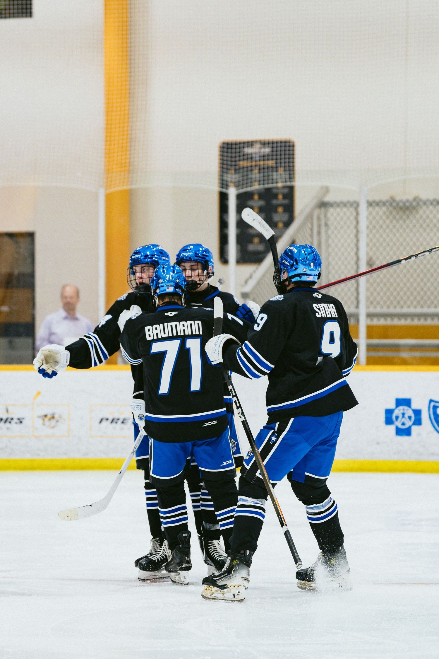 Youth hockey players in black and blue uniforms celebrate together on ice after scoring a goal.